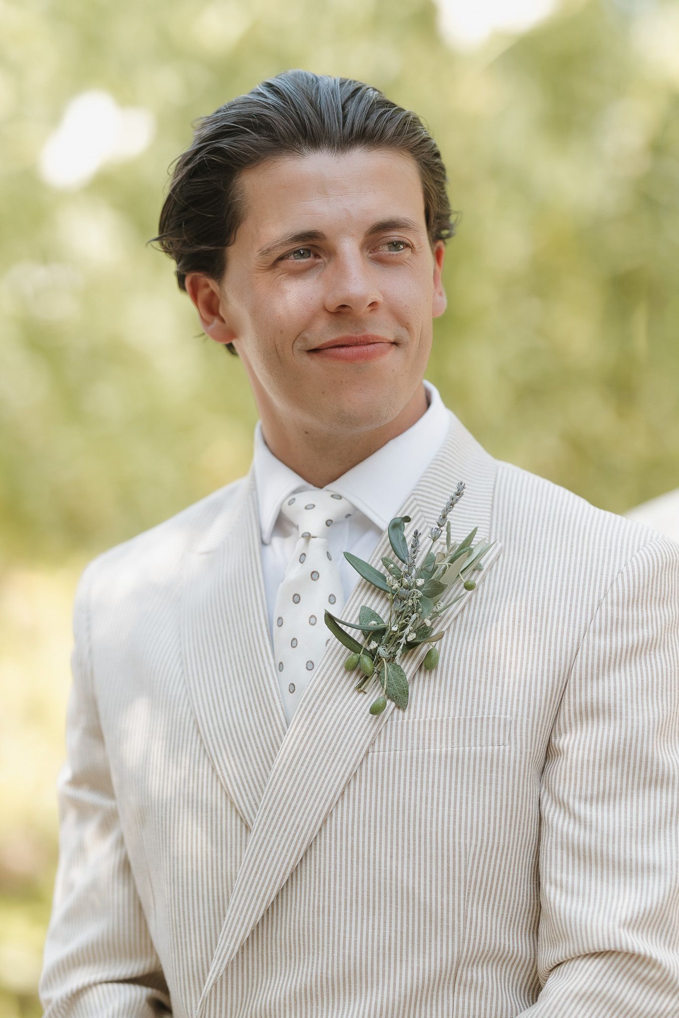 A groom in a striped seersucker suit with a greenery boutonniere and polka dot tie smiles outdoors.