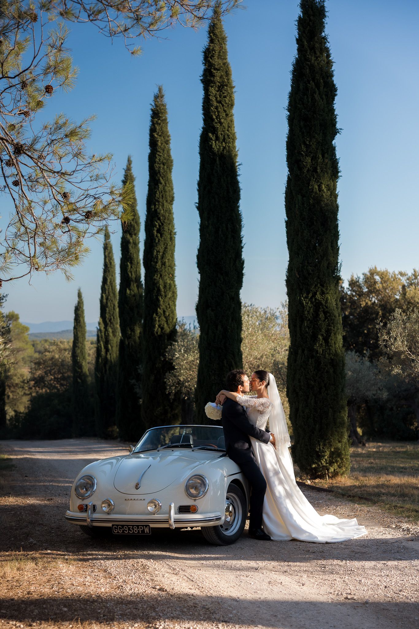 Bride and groom kissing beside a vintage white Porsche 356 on a gravel path lined with tall cypress trees.