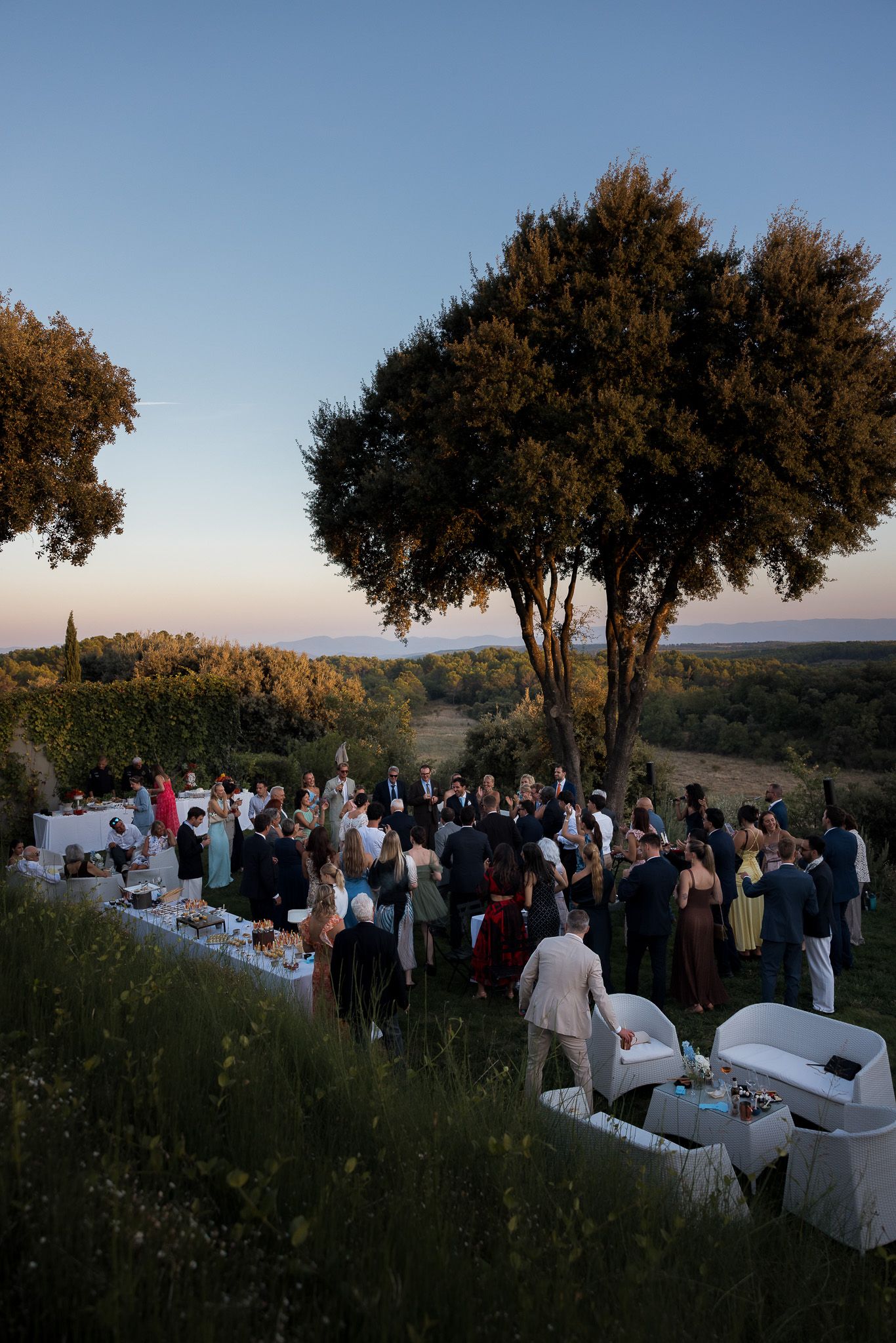 Outdoor wedding reception at sunset with guests gathered around tables on a hillside, surrounded by trees and scenic countryside views.