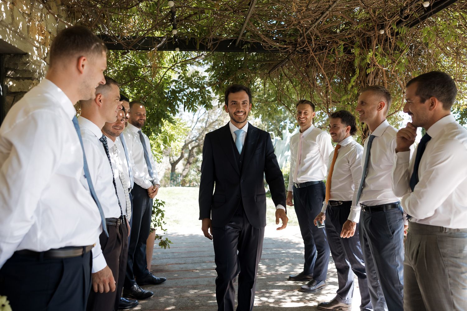 Groom in dark suit greets groomsmen in white shirts and ties outdoors under a vine-covered pergola.