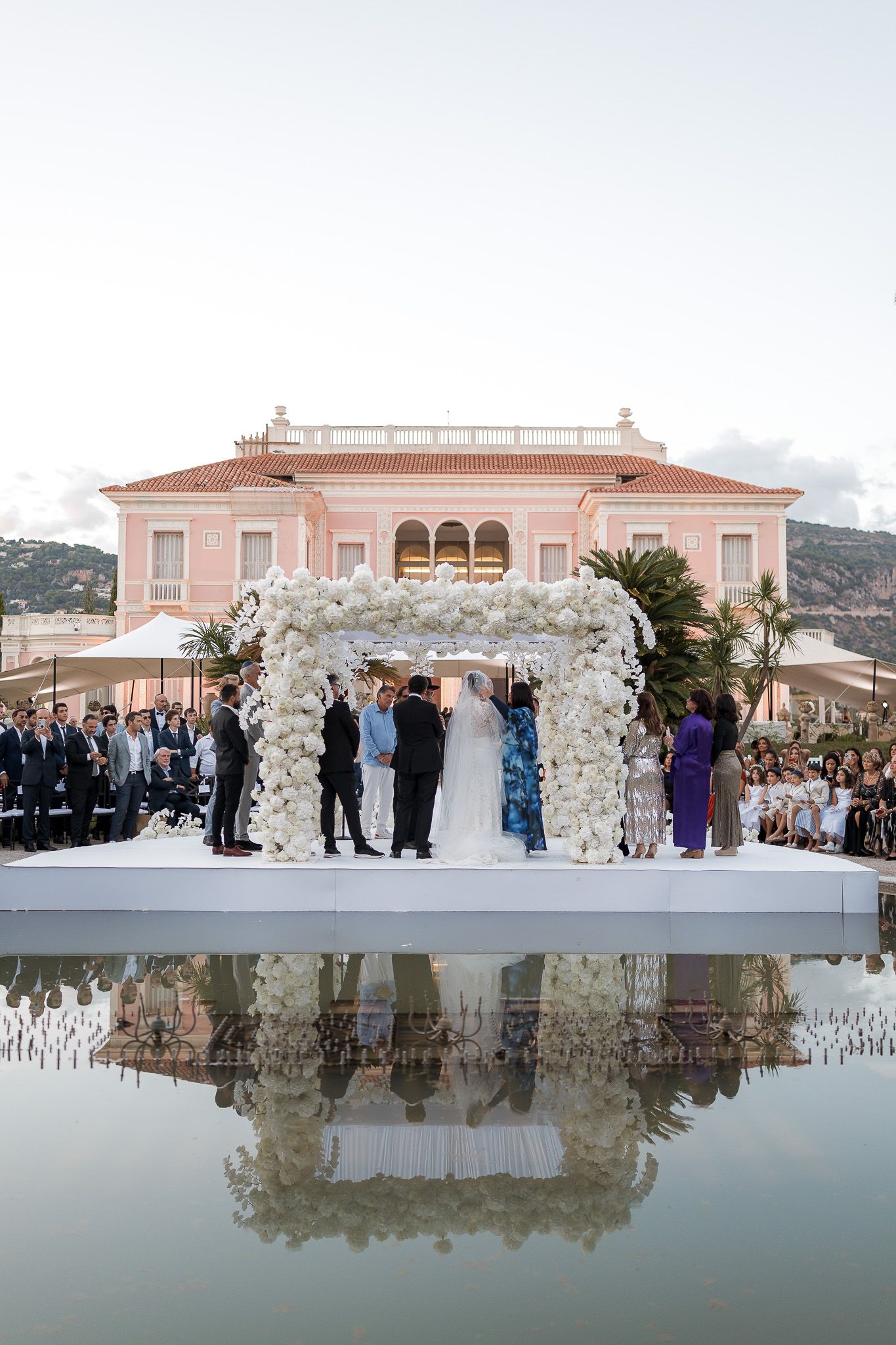 Outdoor wedding ceremony under white floral arch on reflective pool stage with pink villa backdrop.