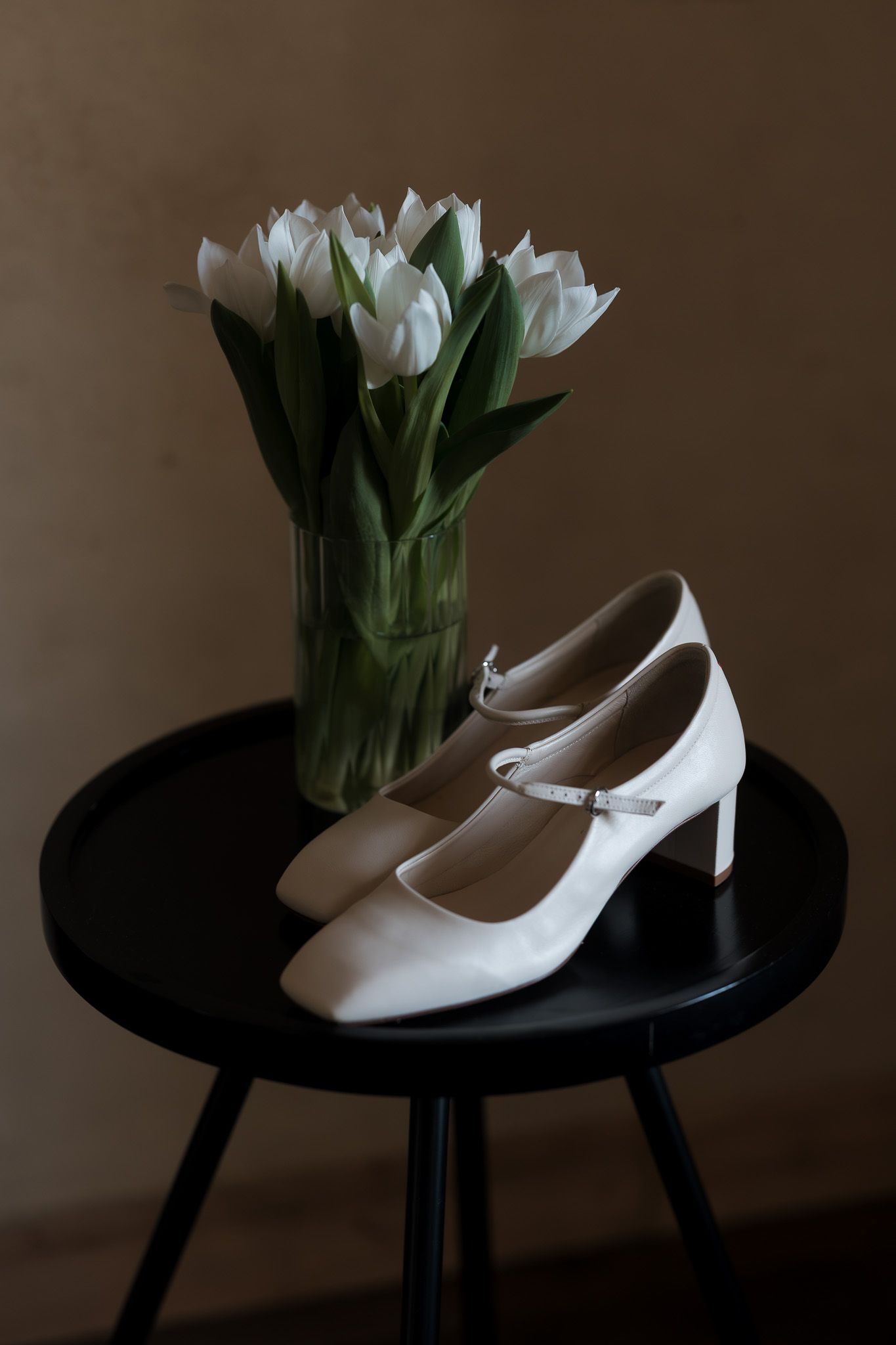 White Mary Jane heels beside a vase of white tulips on a black side table.