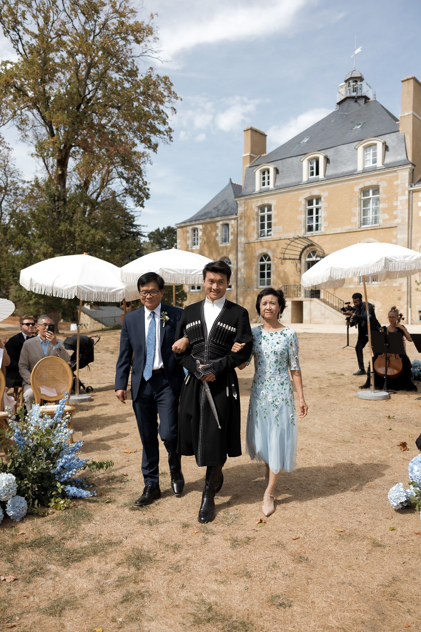 Groom in traditional attire walks with family outside a French château during an outdoor wedding ceremony.