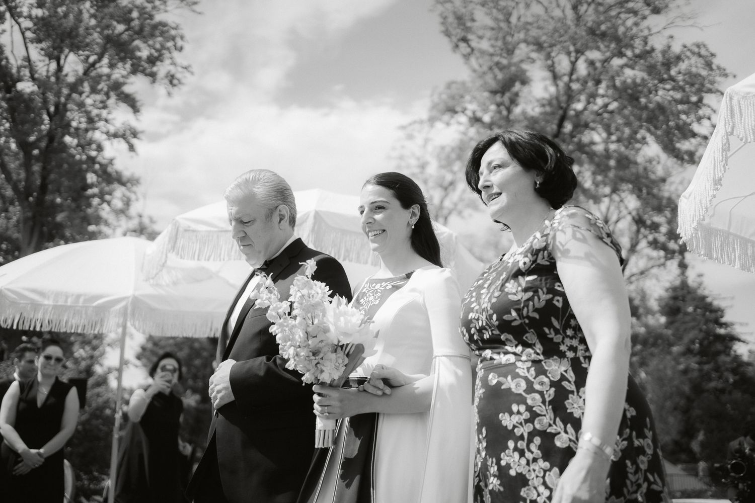 Black and white wedding photo of bride walking down aisle with parents, holding bouquet, guests and umbrellas in background.