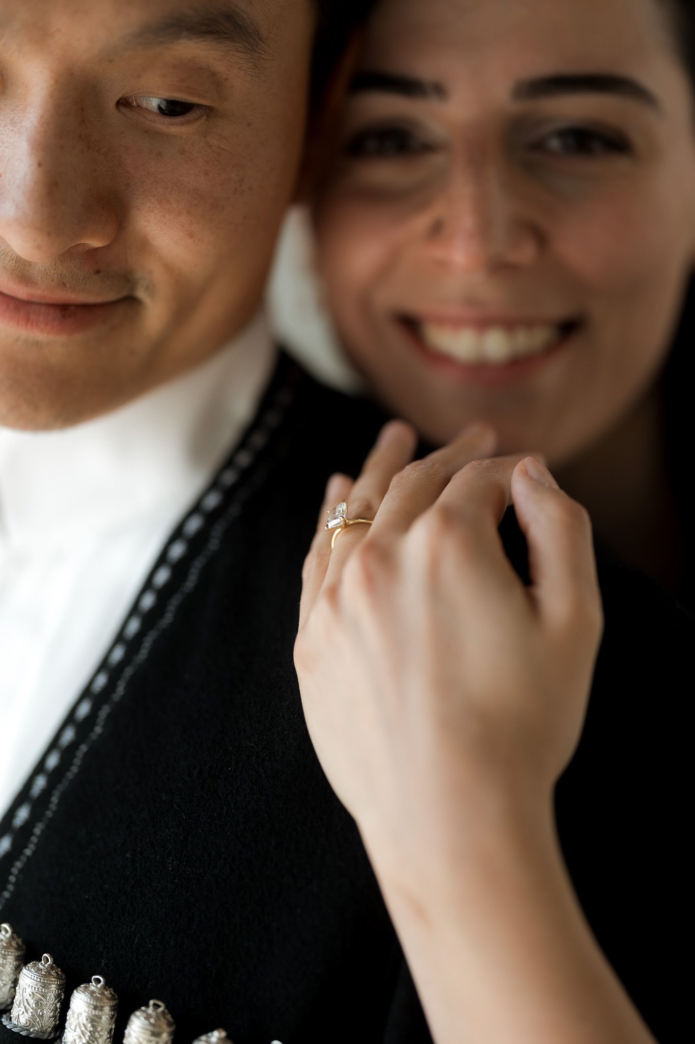 Couple smiling and showing off engagement ring, man in black traditional outfit, woman displaying gold diamond ring on finger.