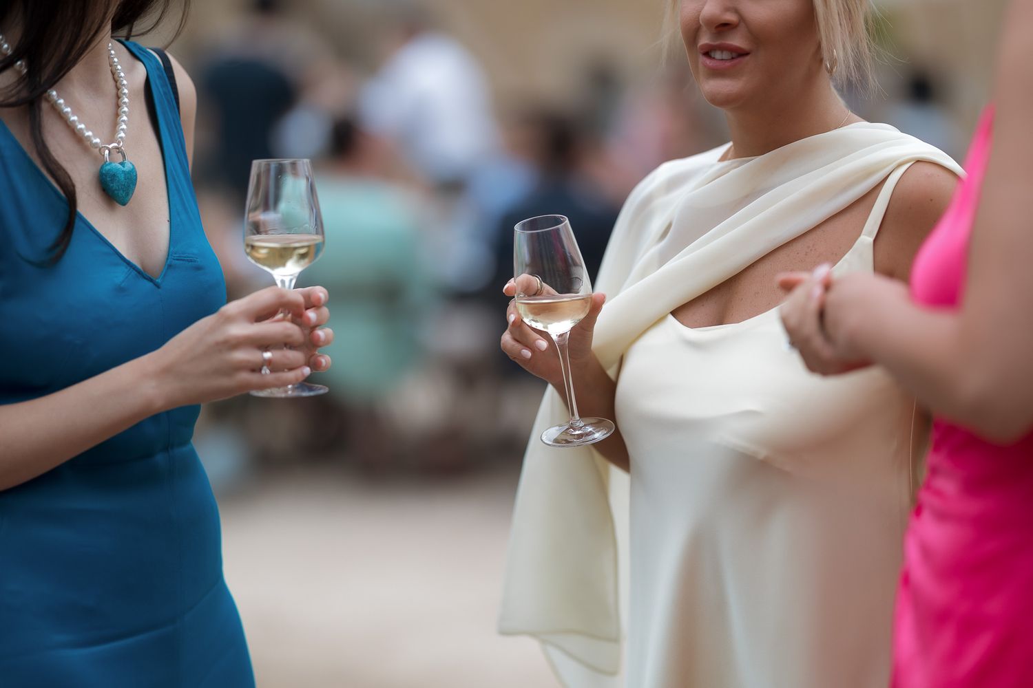 Women in elegant dresses holding wine glasses at a sophisticated outdoor social event or wedding reception.