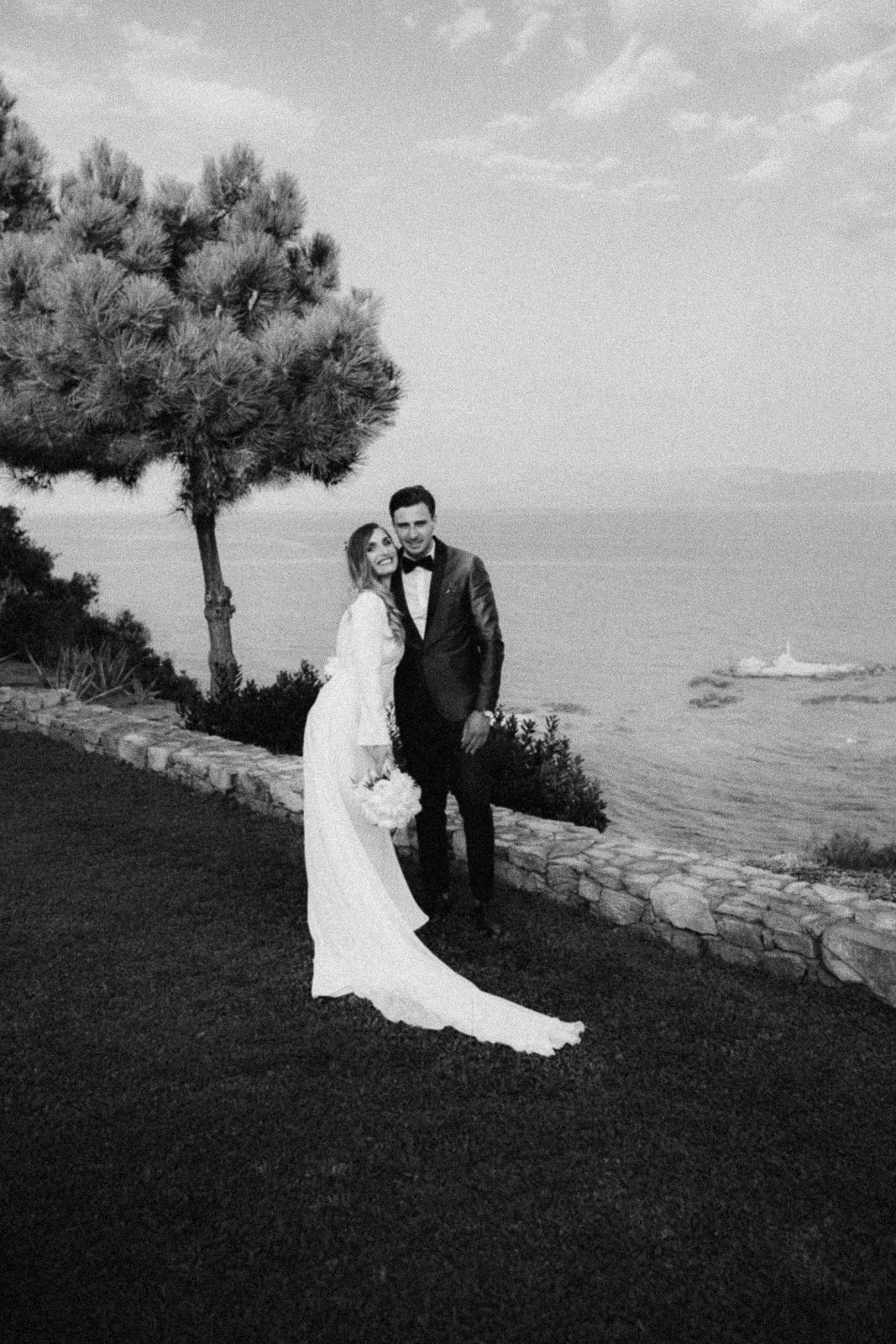 A black and white wedding photo shows a couple embracing on a coastal cliff overlooking the ocean with a pine tree.
