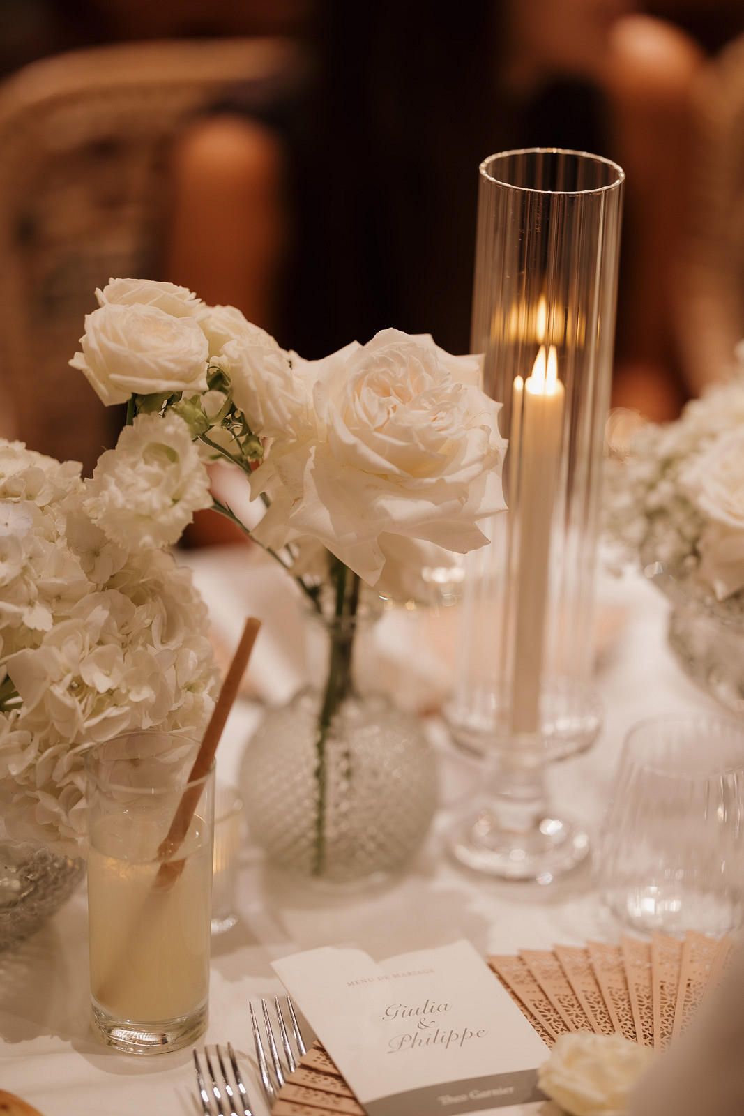 Elegant white roses and hydrangeas in glass vases adorn a wedding reception table with glowing candles and place cards.