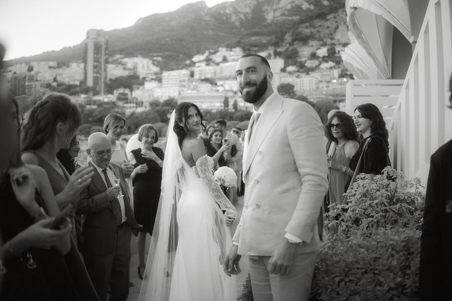 A black and white wedding photograph showing guests gathered outdoors at a scenic hillside venue.
