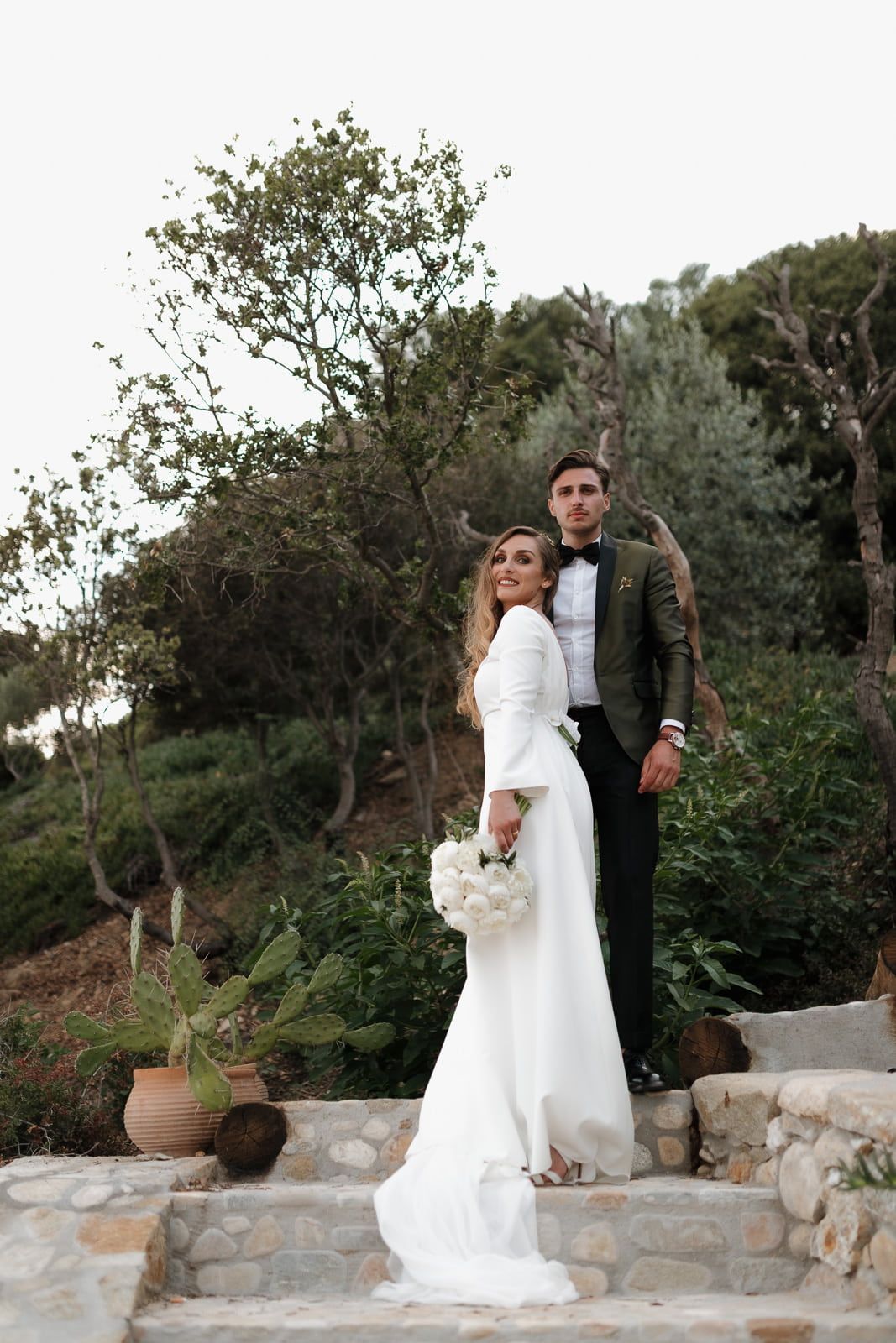 Romantic wedding couple embraces on stone terrace surrounded by olive trees at sunset in Mediterranean setting.