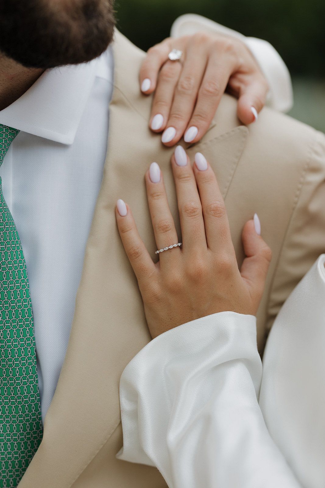 Close-up view of hands wearing white nail polish and wedding rings against beige and white formal attire.