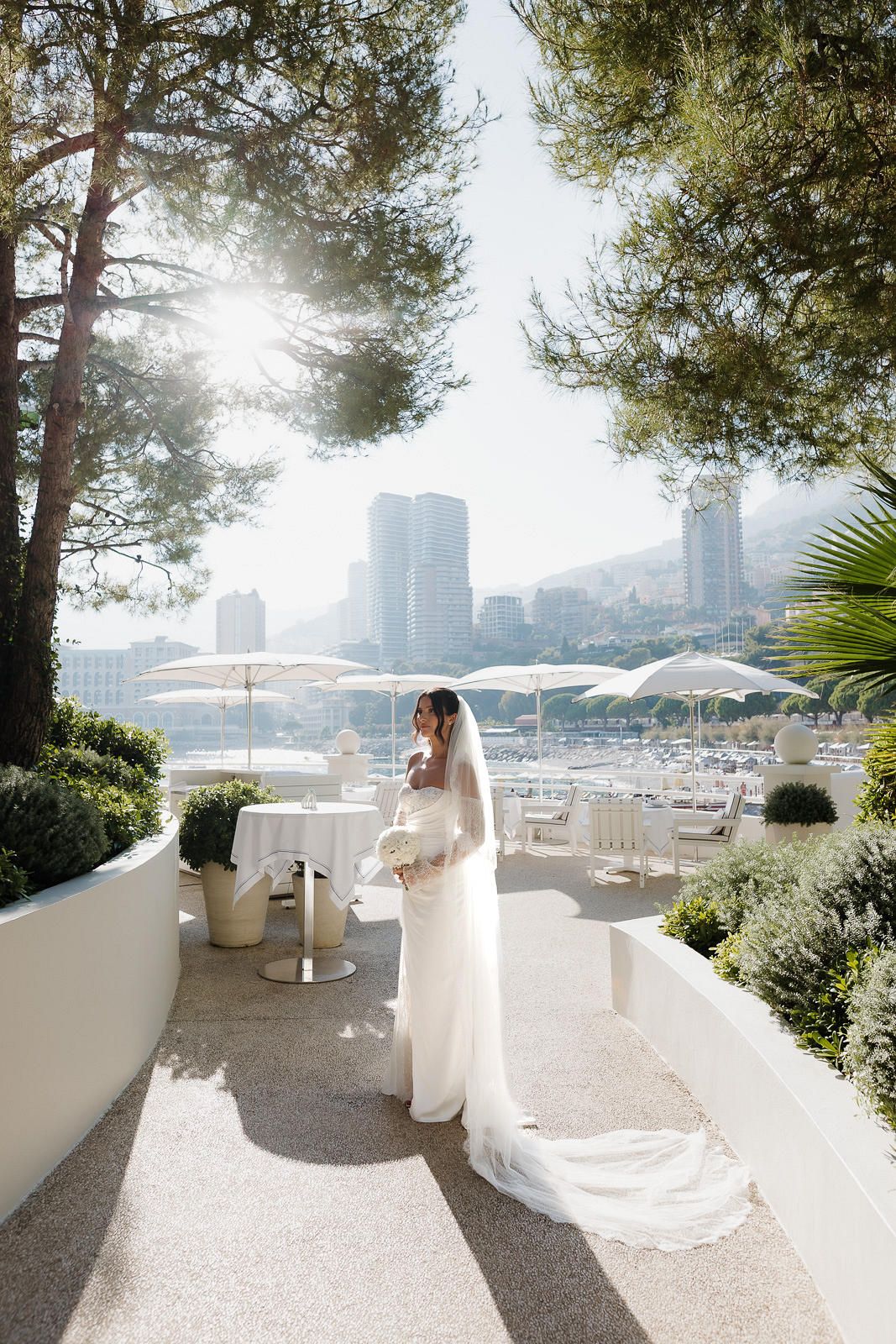 A sunlit outdoor terrace with white umbrellas and chairs overlooking a city skyline through lush green trees.