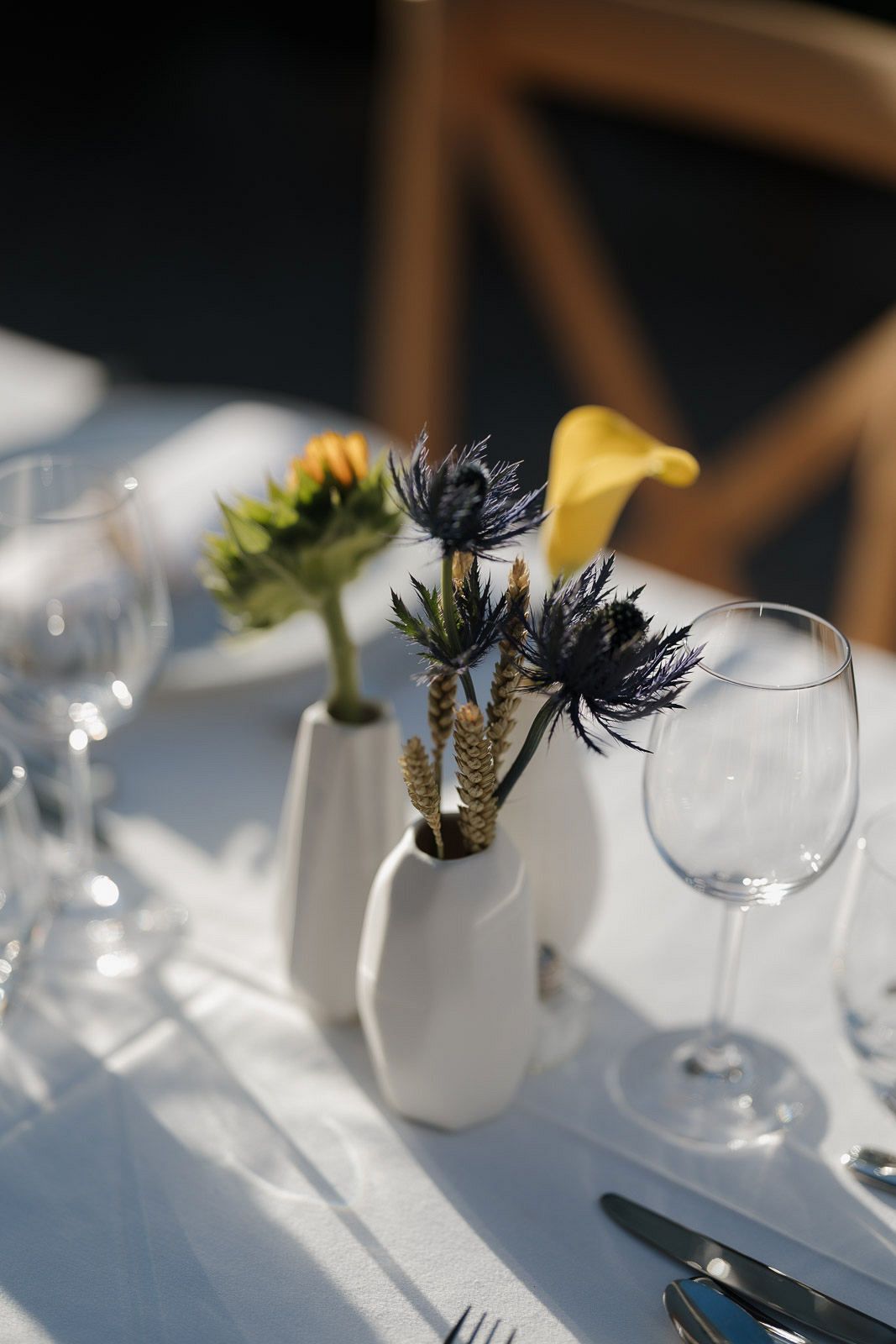 Elegant table setting with white vases, wine glasses and delicate thistle flowers in natural sunlight.