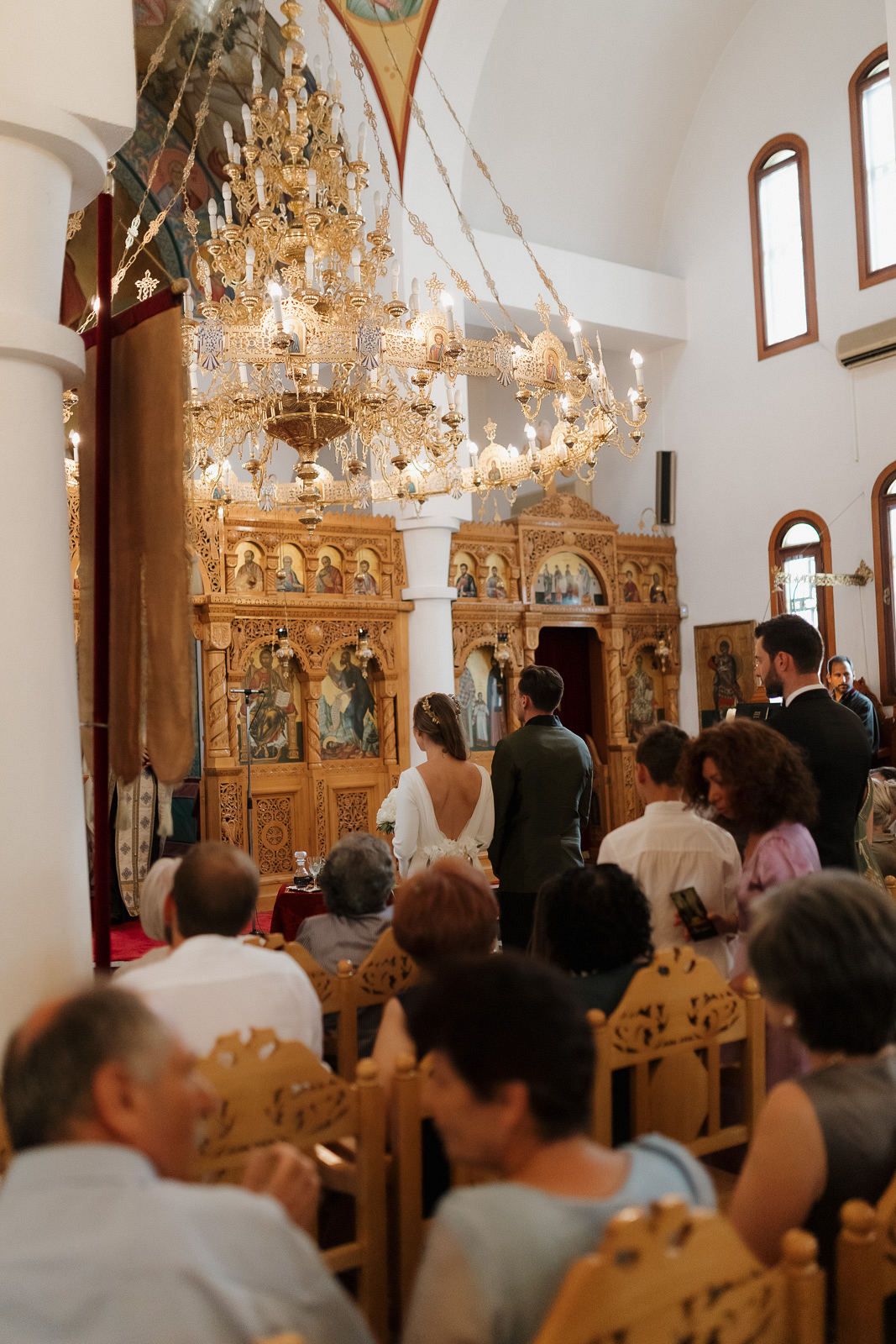 Guests gather inside an ornate church with chandeliers and wooden pews during a wedding ceremony.