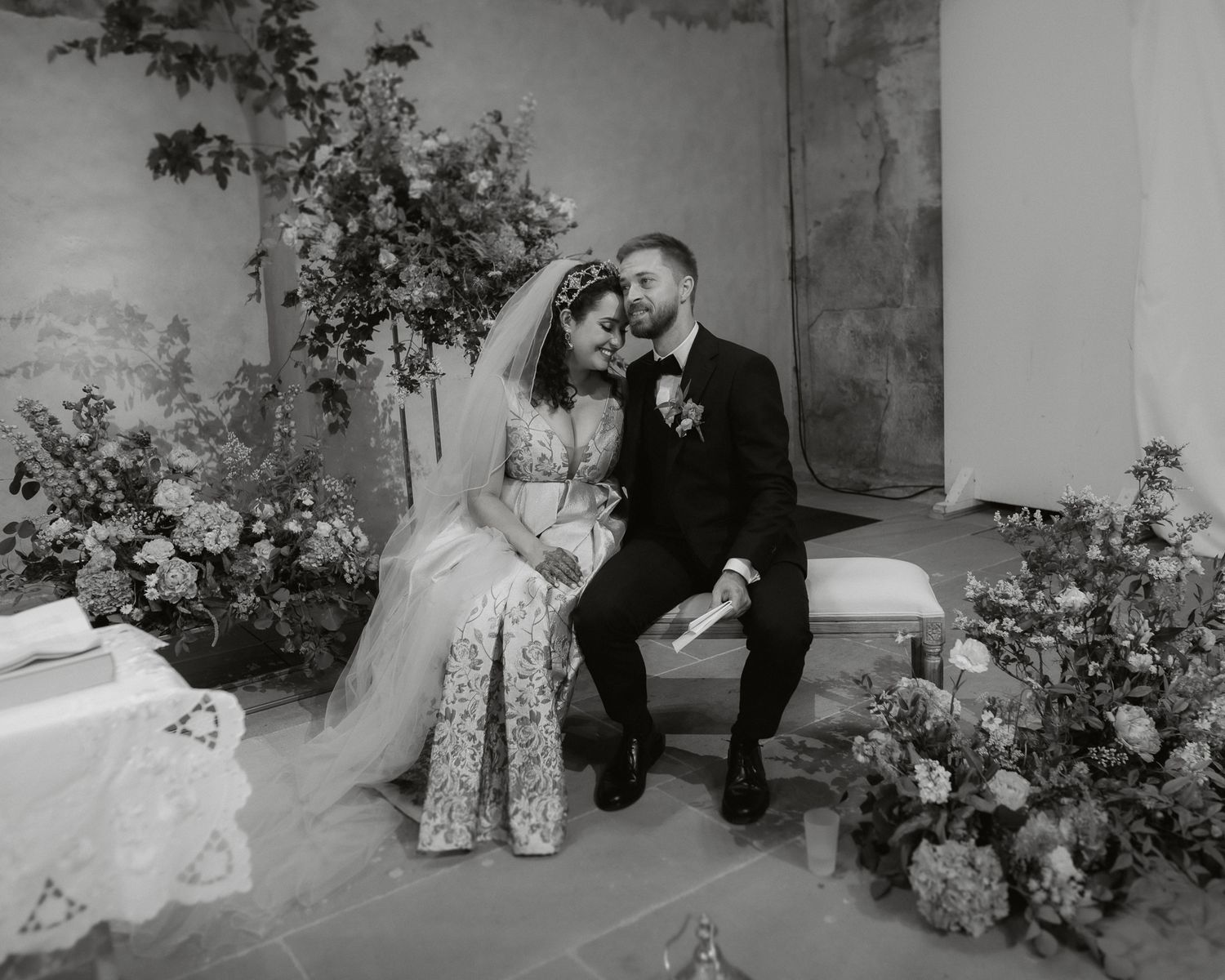 A black and white wedding portrait with couple seated among floral arrangements.