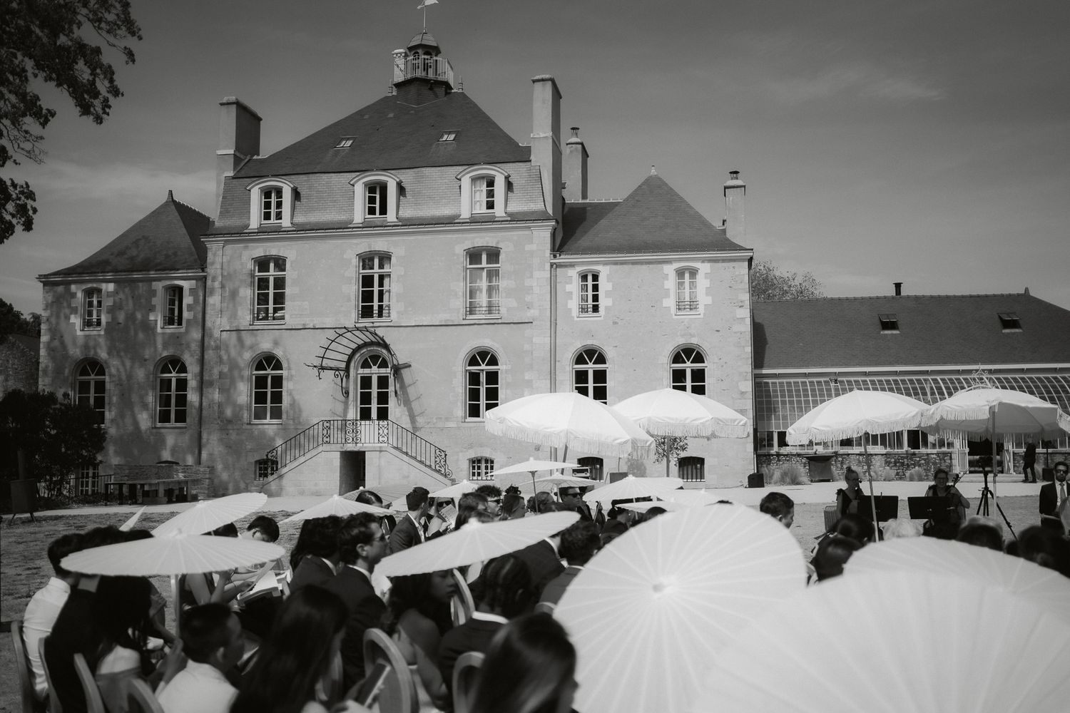 Black and white photo of wedding guests holding white parasols outside an elegant French chateau.