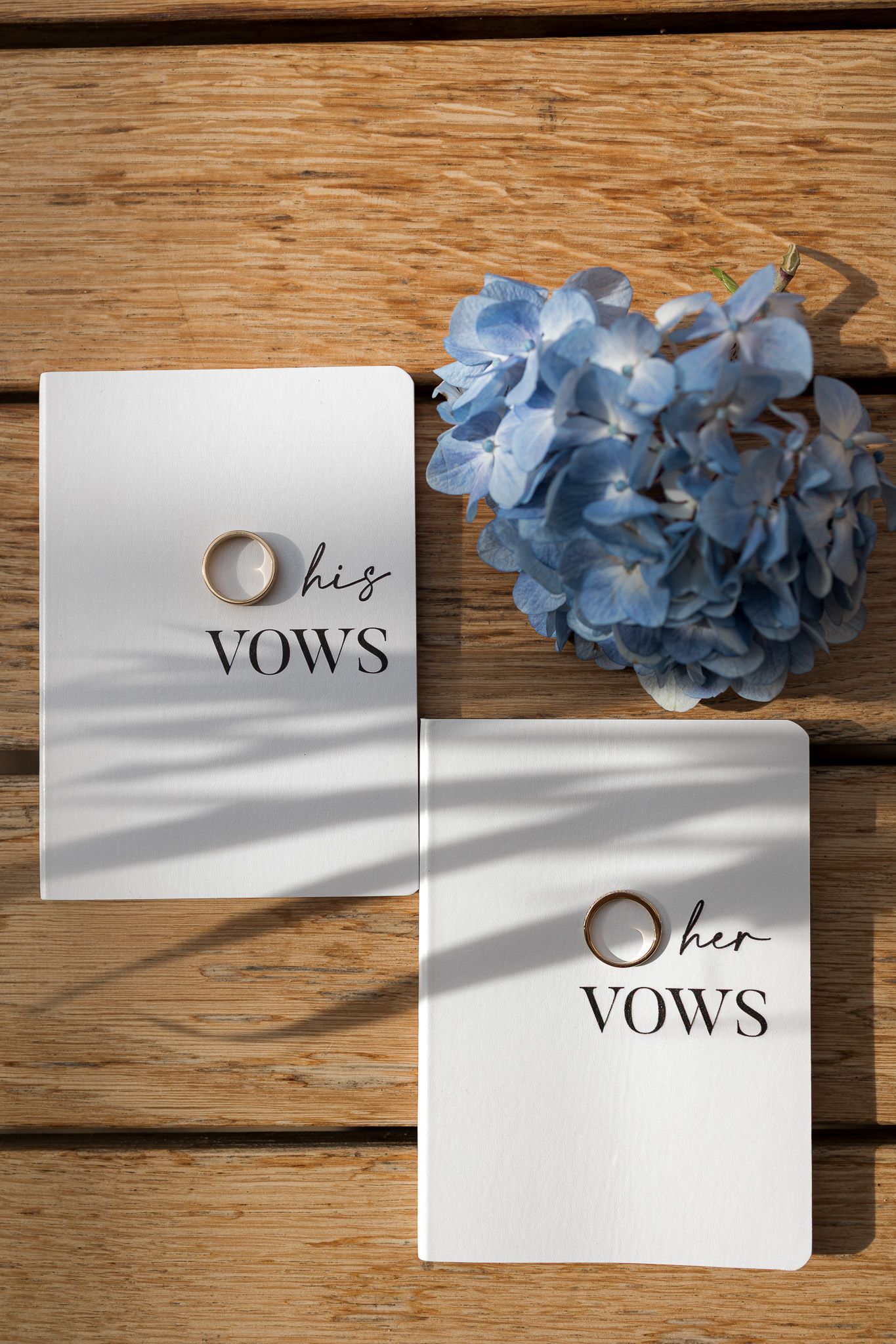 His and her vows booklets with gold wedding rings and blue hydrangea flowers on rustic wooden background.