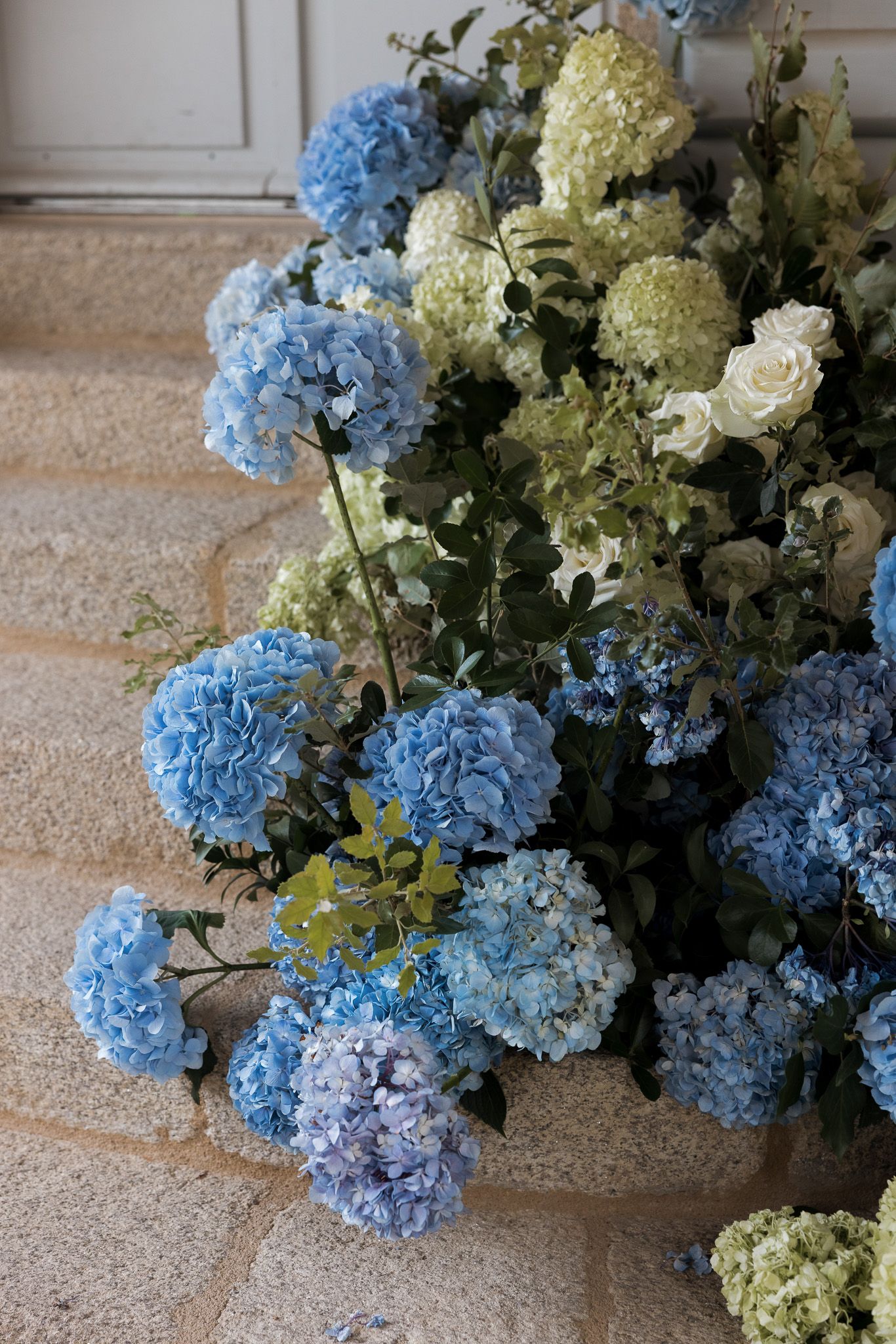 Blue and white hydrangeas with roses arranged in a lush floral display on stone steps near a door.