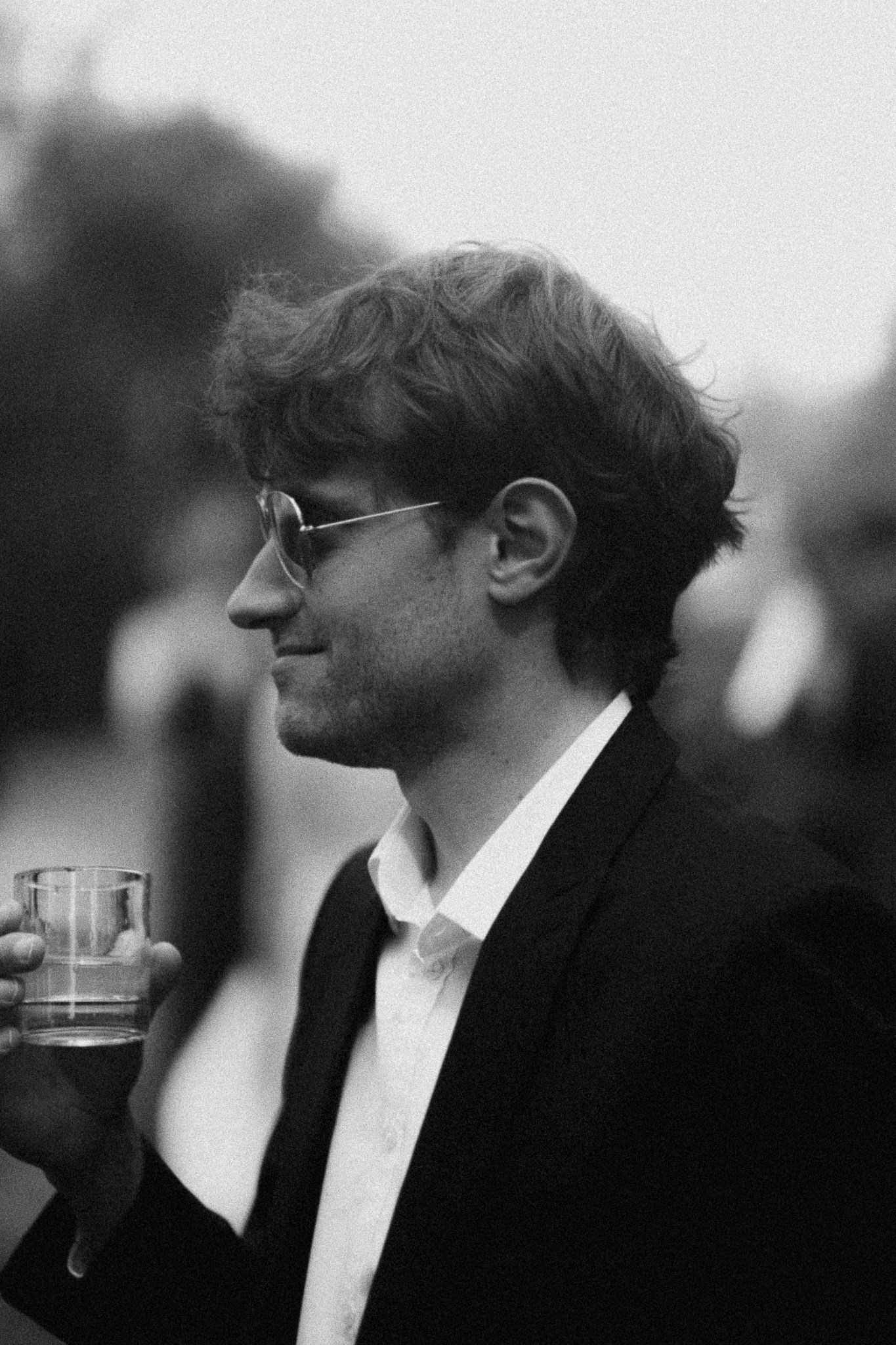 Black and white photo of a young man in a suit wearing glasses, holding a glass drink outdoors.