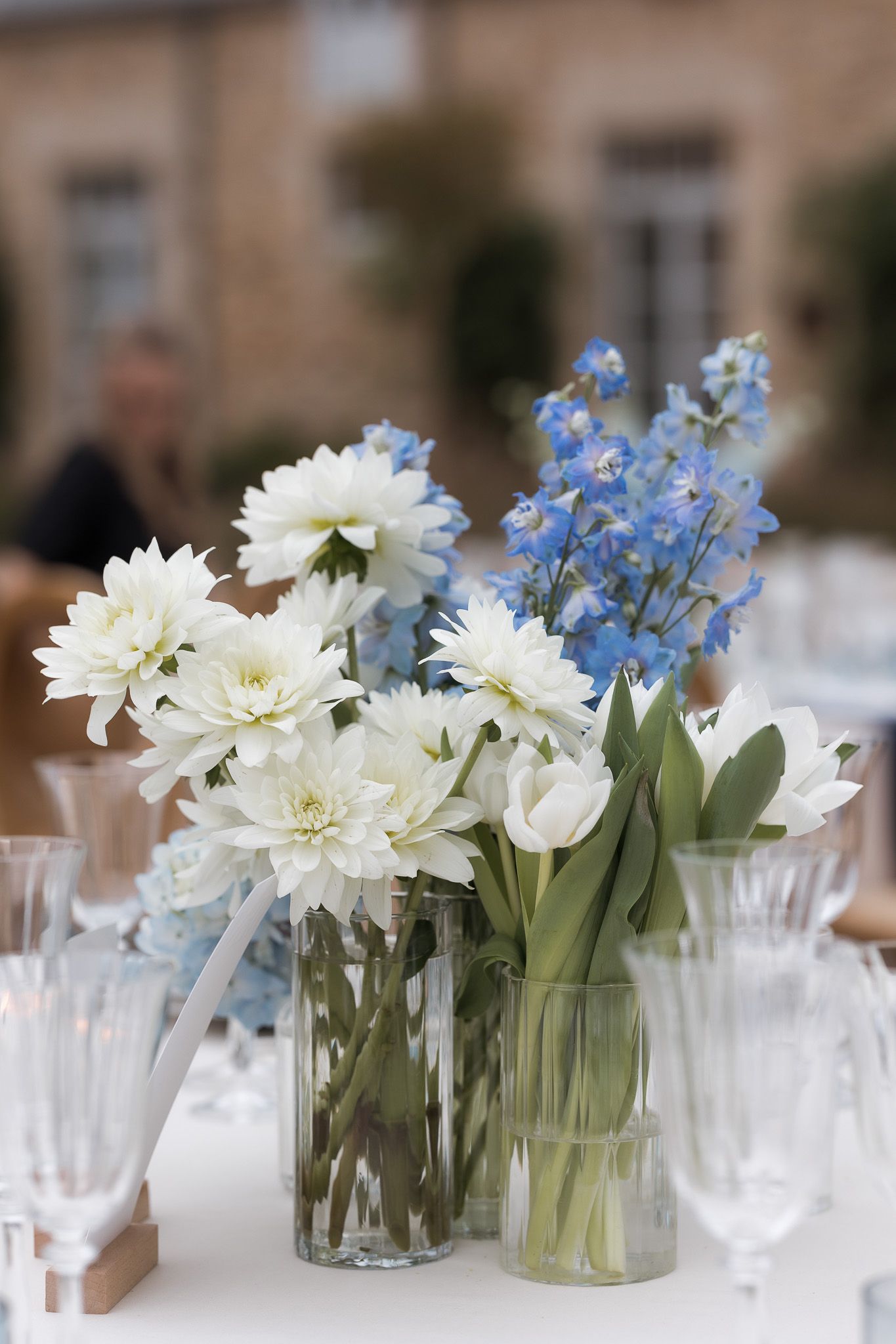 Elegant table centerpiece with white dahlias, blue delphiniums, and tulips in clear glass vases at an outdoor event.