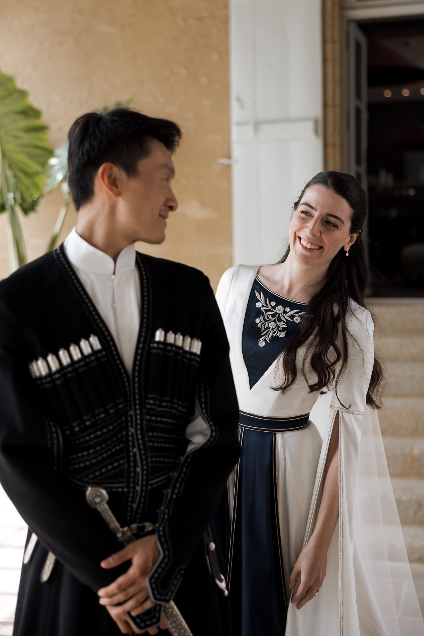 Couple in traditional cultural attire smiling at each other, man holding ceremonial sword, woman in elegant white and navy dress.