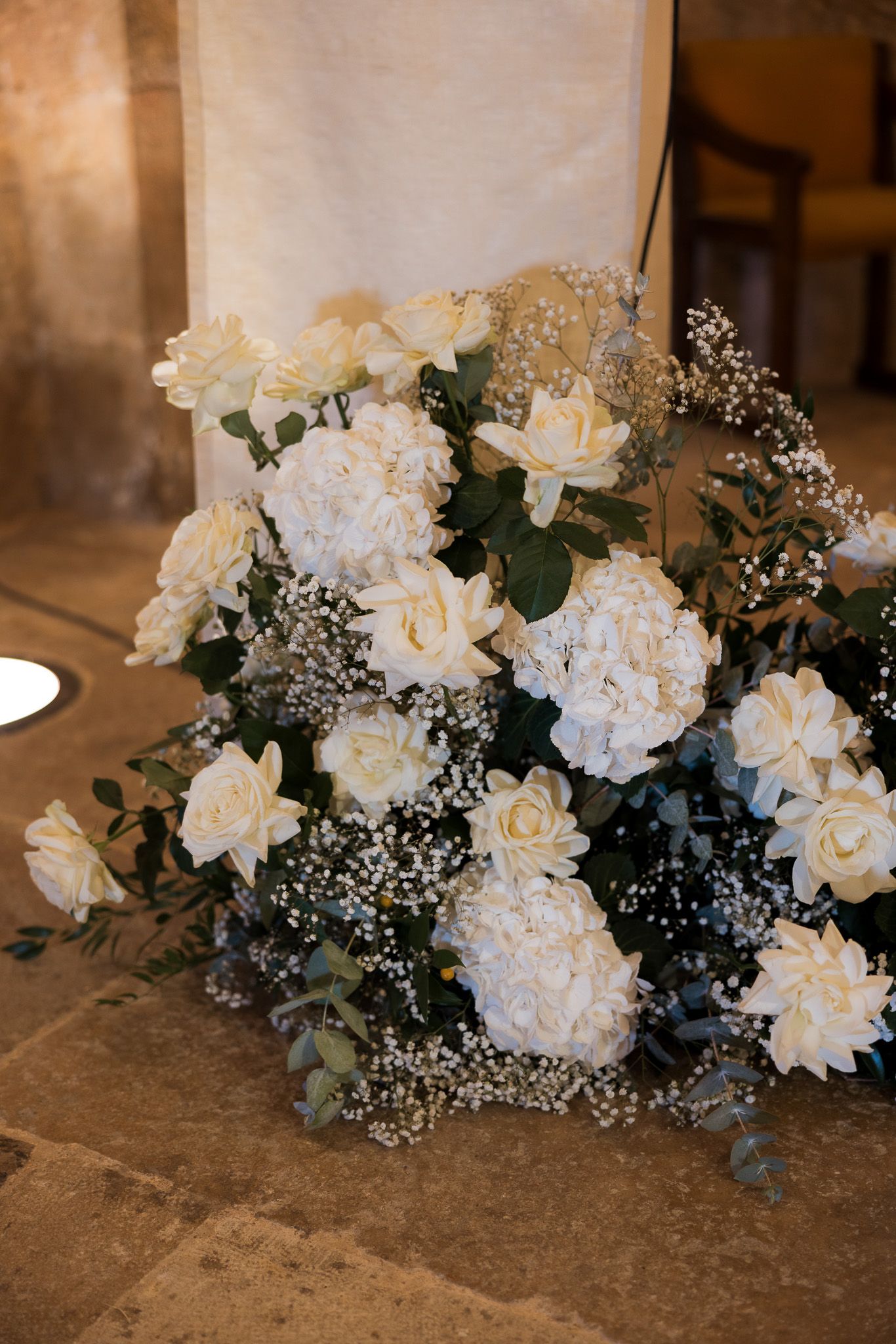 Elegant white floral arrangement with roses, hydrangeas, and baby's breath on a stone floor.