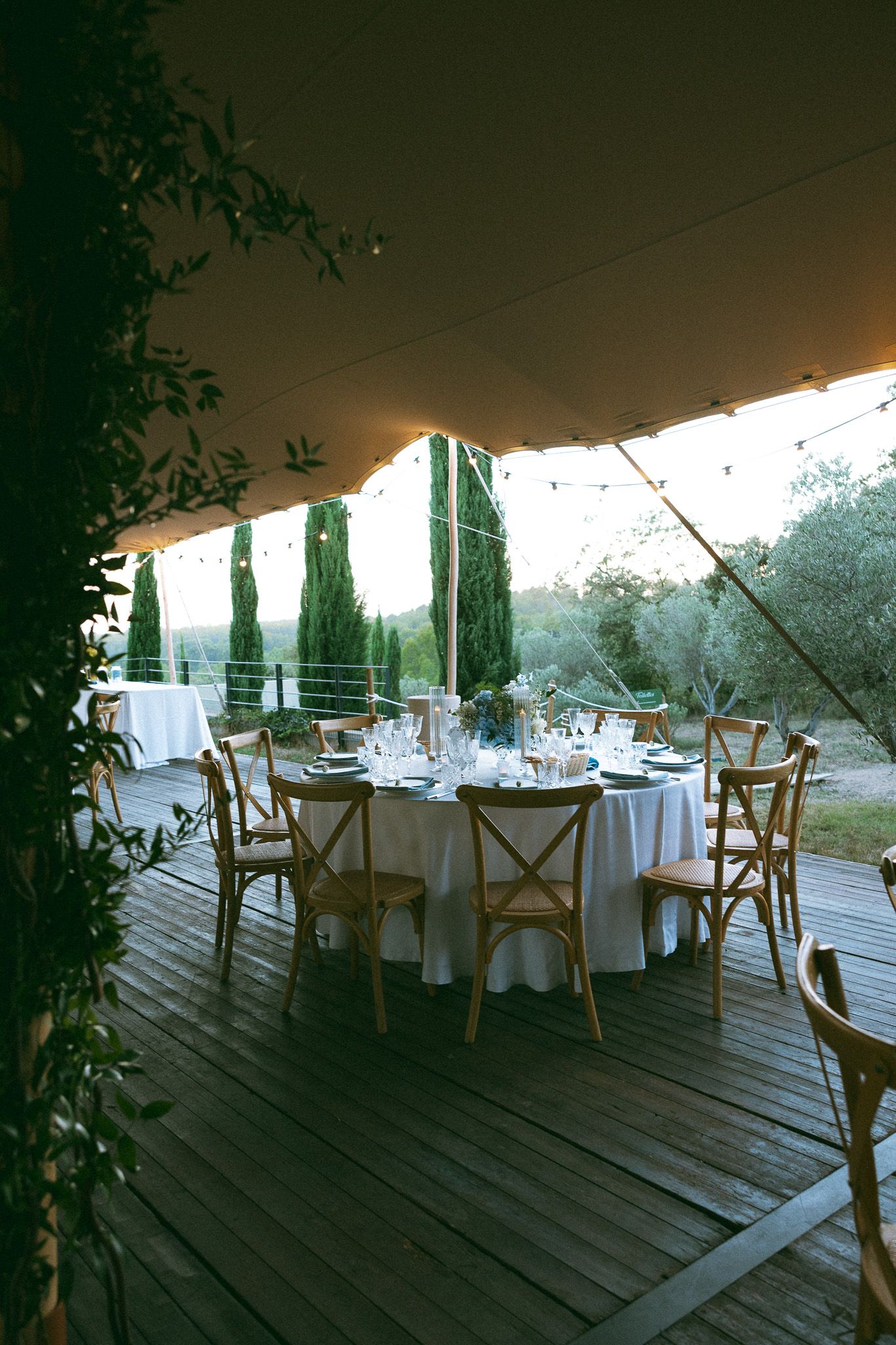 Elegant outdoor wedding reception under a tent on a wooden deck with round tables, cross-back chairs, and cypress trees.