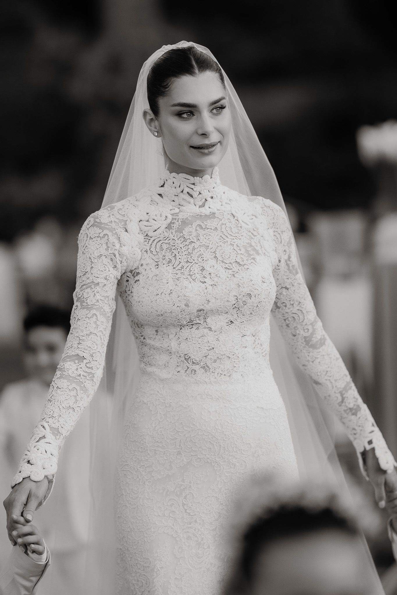 Elegant bride in long-sleeve lace wedding gown with high neck and veil, black and white photo.