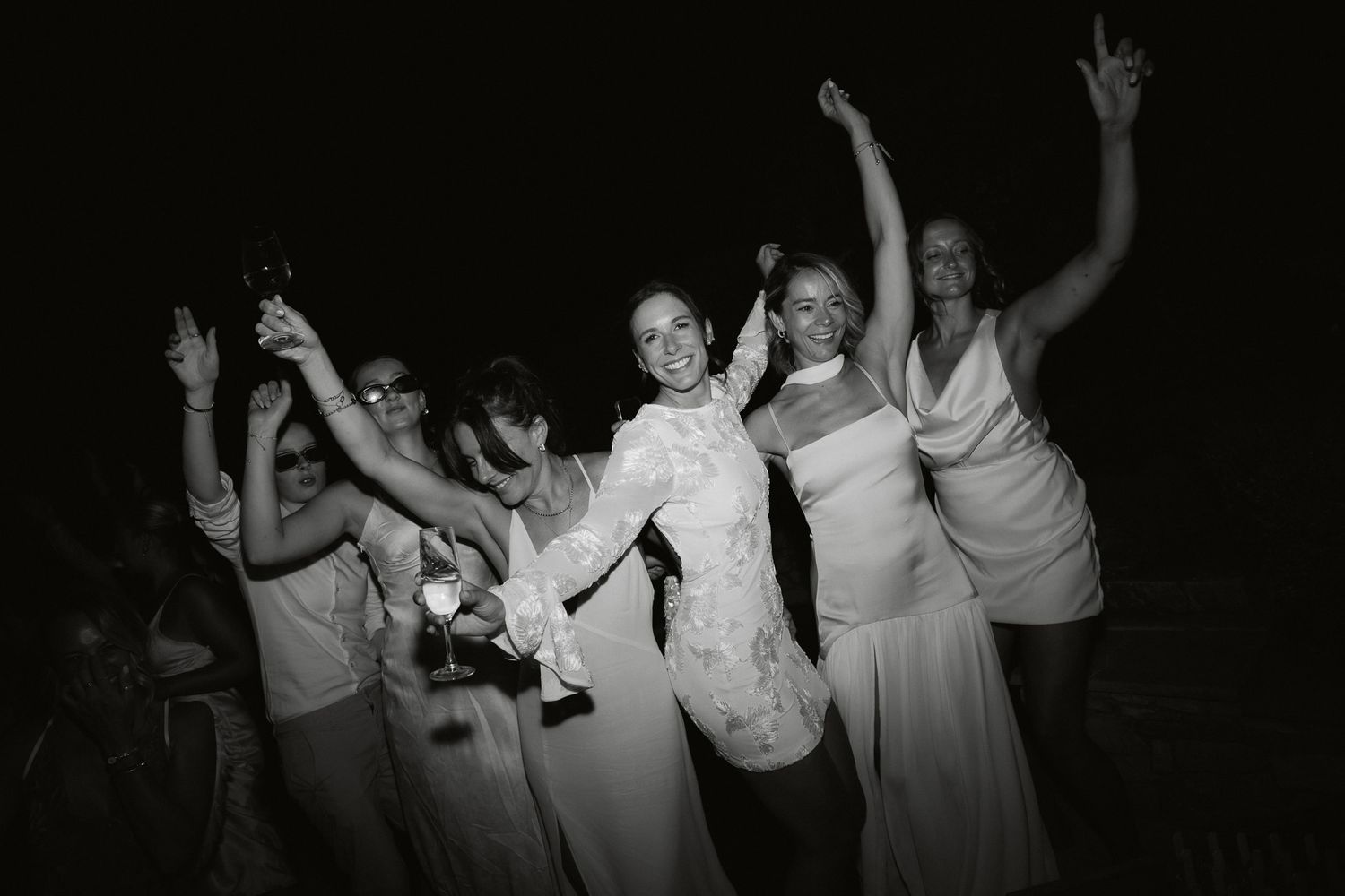 Black and white photo of a bride in lace gown celebrating with bridesmaids in white dresses at a wedding reception.
