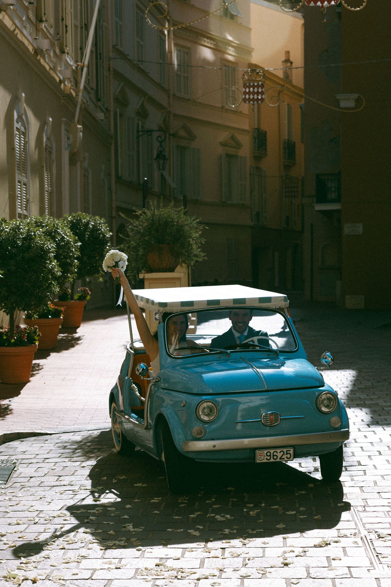 Vintage blue Fiat 500 parked on cobblestone street in Italian town with potted plants and sunlit buildings.