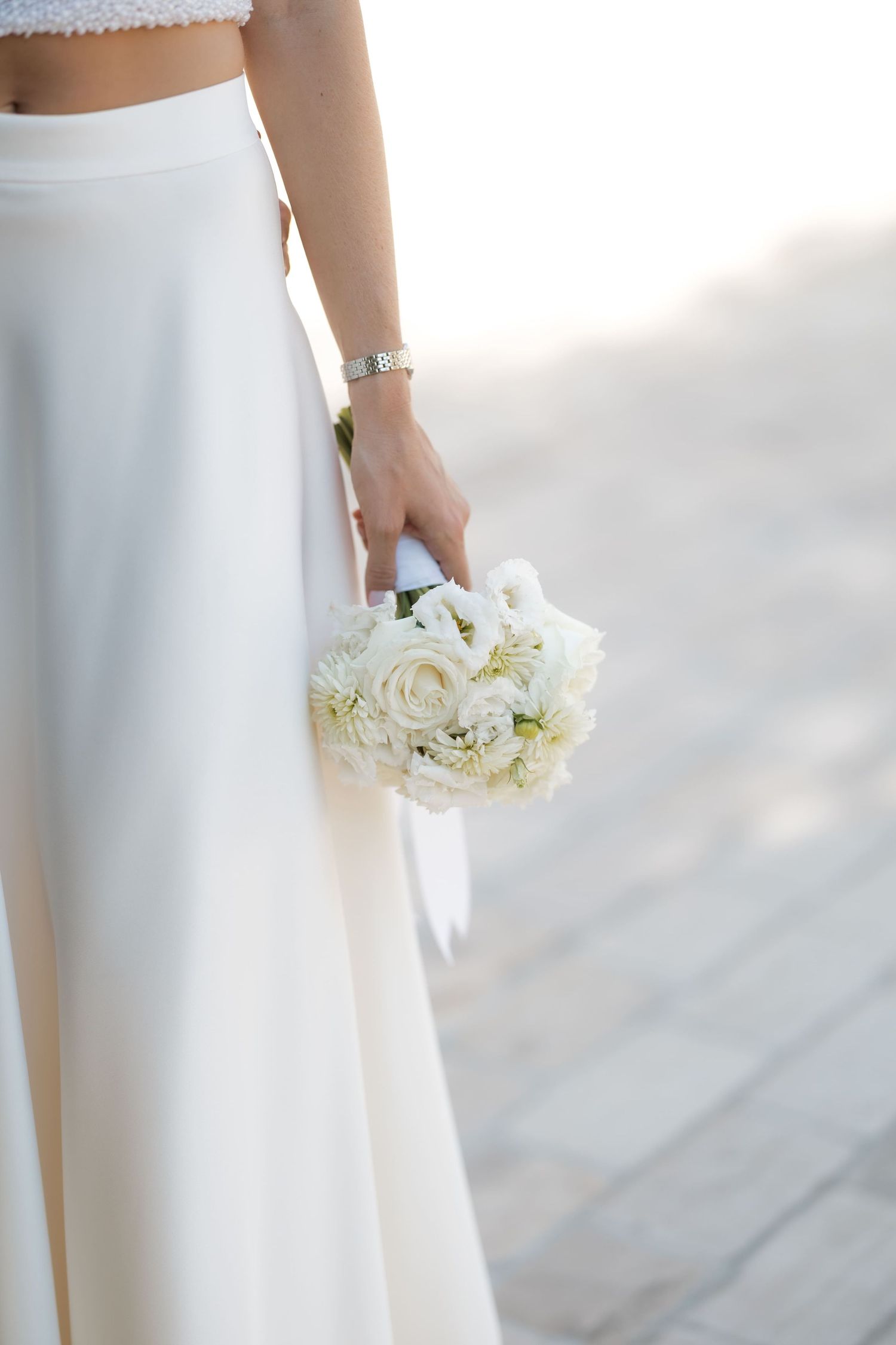 Bride in white two-piece gown holding elegant white rose bouquet at her side, wearing a delicate bracelet.