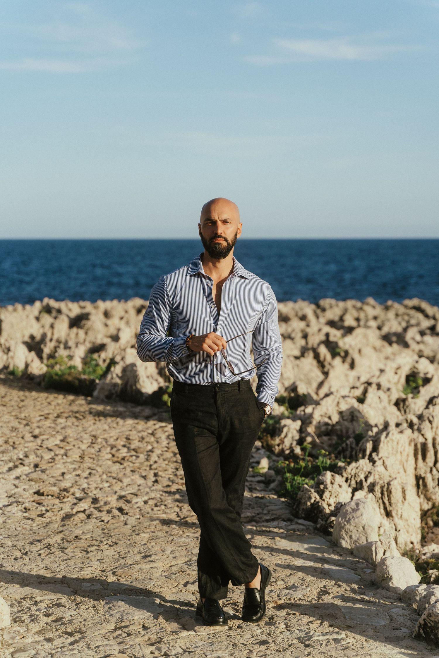 Bald bearded man in blue shirt and black trousers walking on rocky coastal path with ocean in background.