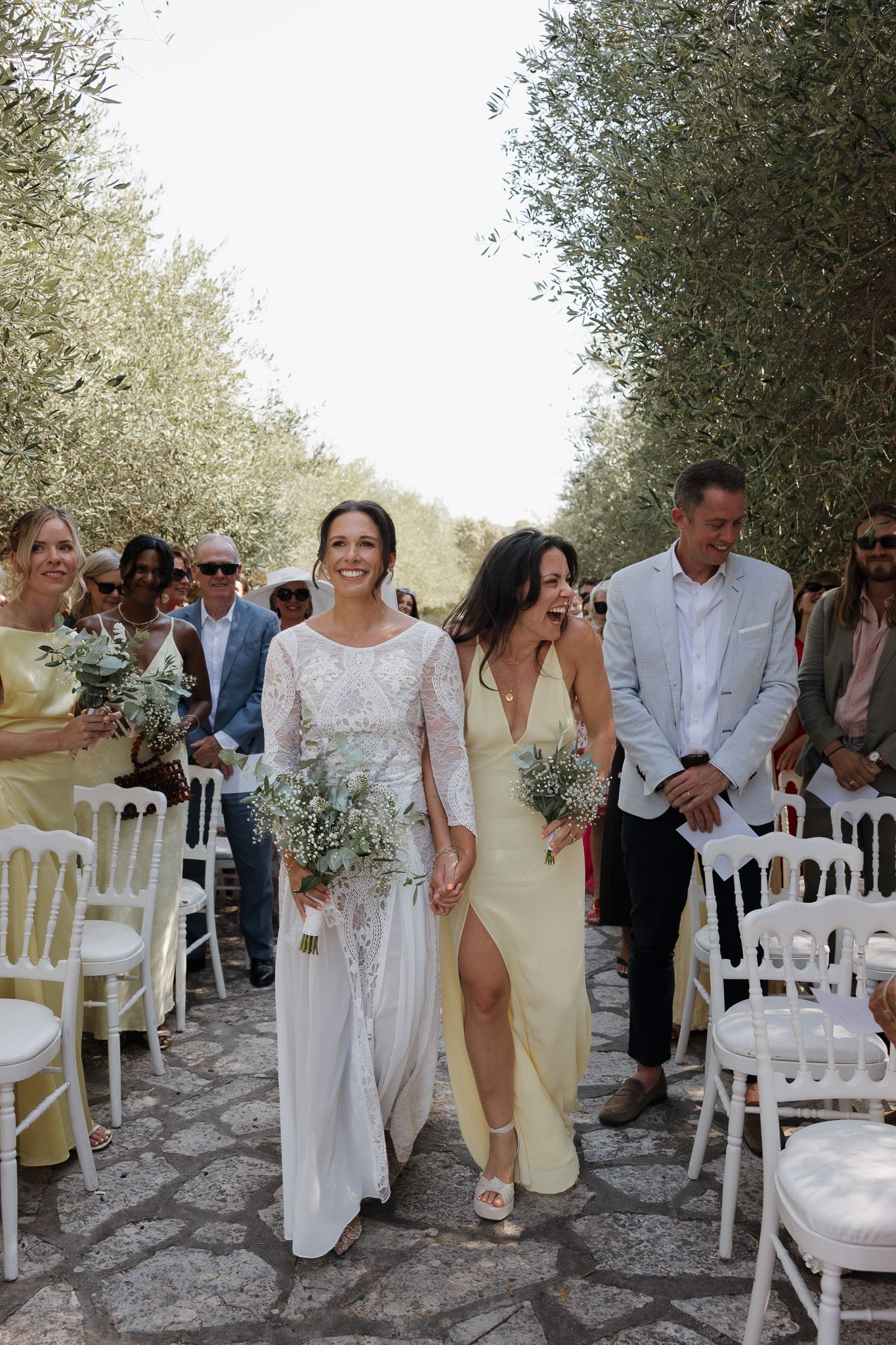 Bride walks down olive tree-lined aisle with bridesmaid in yellow dress holding bouquet at outdoor wedding ceremony.