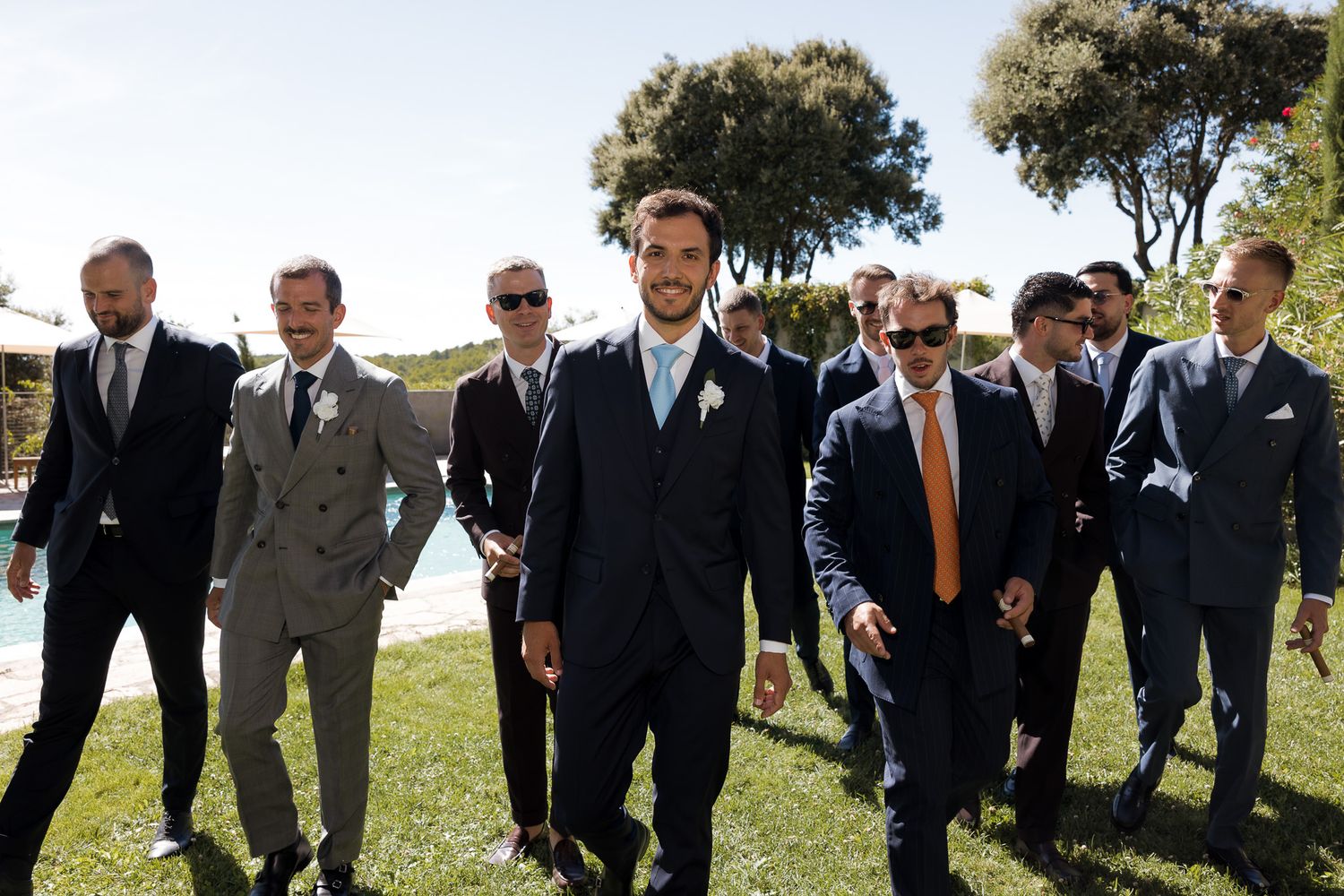 Groom and groomsmen in navy and grey suits walking outdoors at a sunny wedding venue.