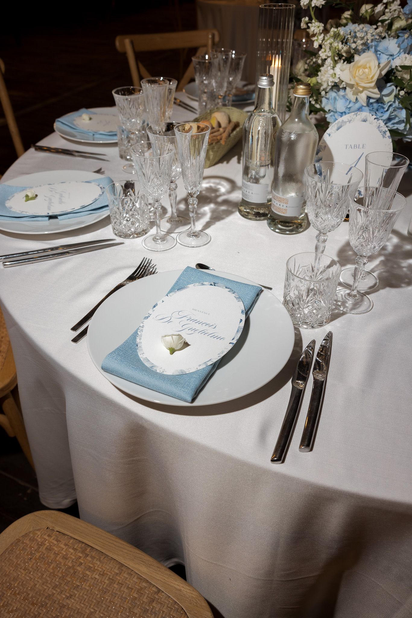 Elegant wedding reception table with crystal glassware, blue napkins, white florals, and personalized place cards on white linen.