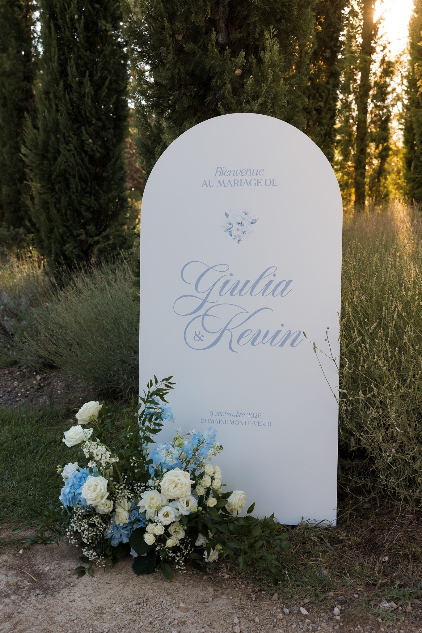 White arched wedding welcome sign for Giulia and Kevin, adorned with blue and white floral arrangement, set outdoors.