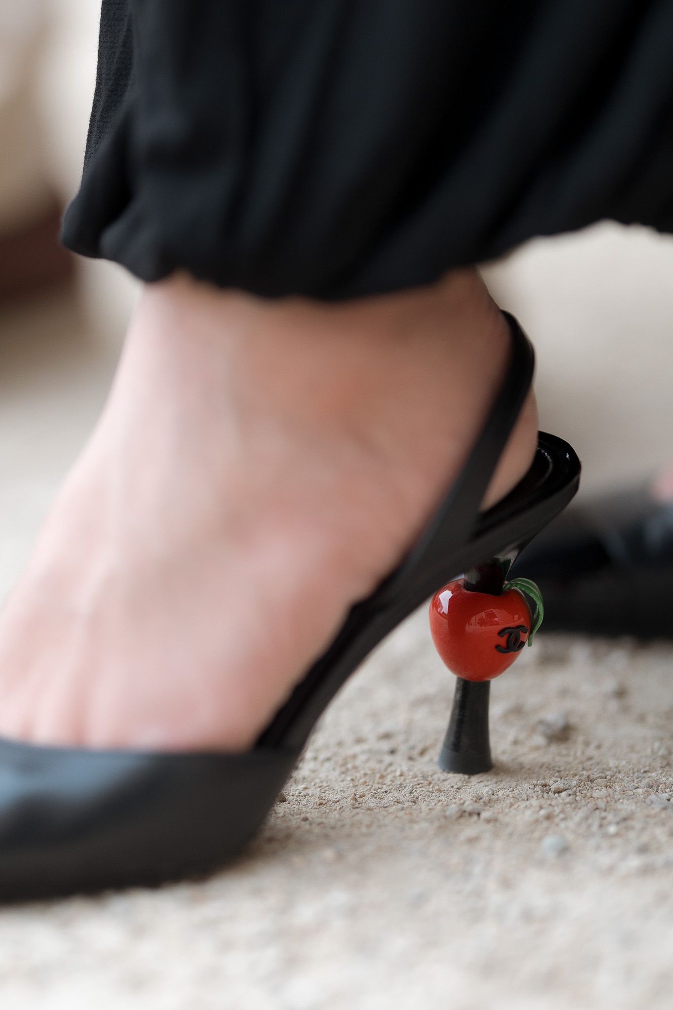 Black slingback stiletto heels with a decorative red apple-shaped heel detail, worn with black trousers.