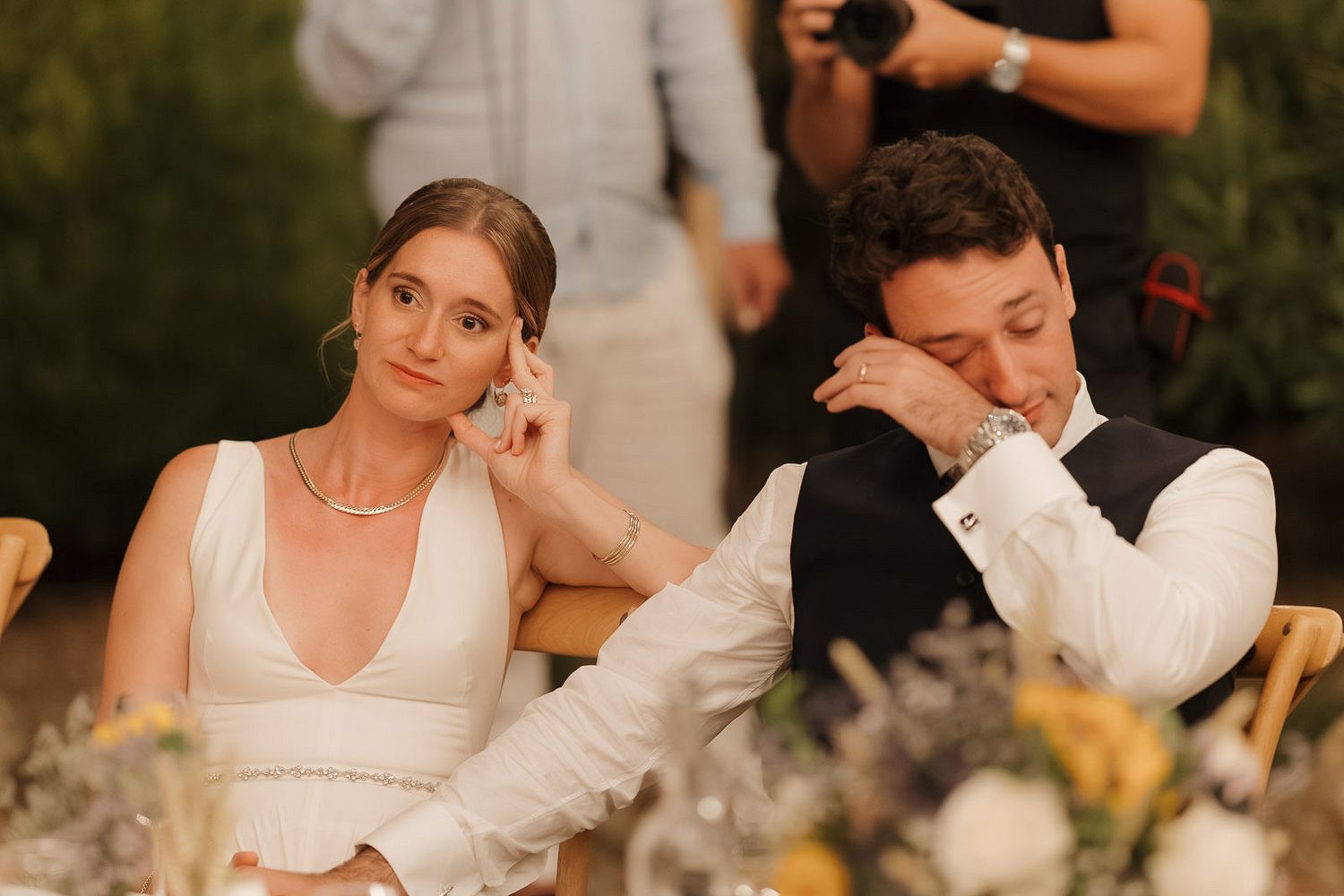 A couple sits at their wedding reception table, with one appearing tired while the other looks on with amusement.