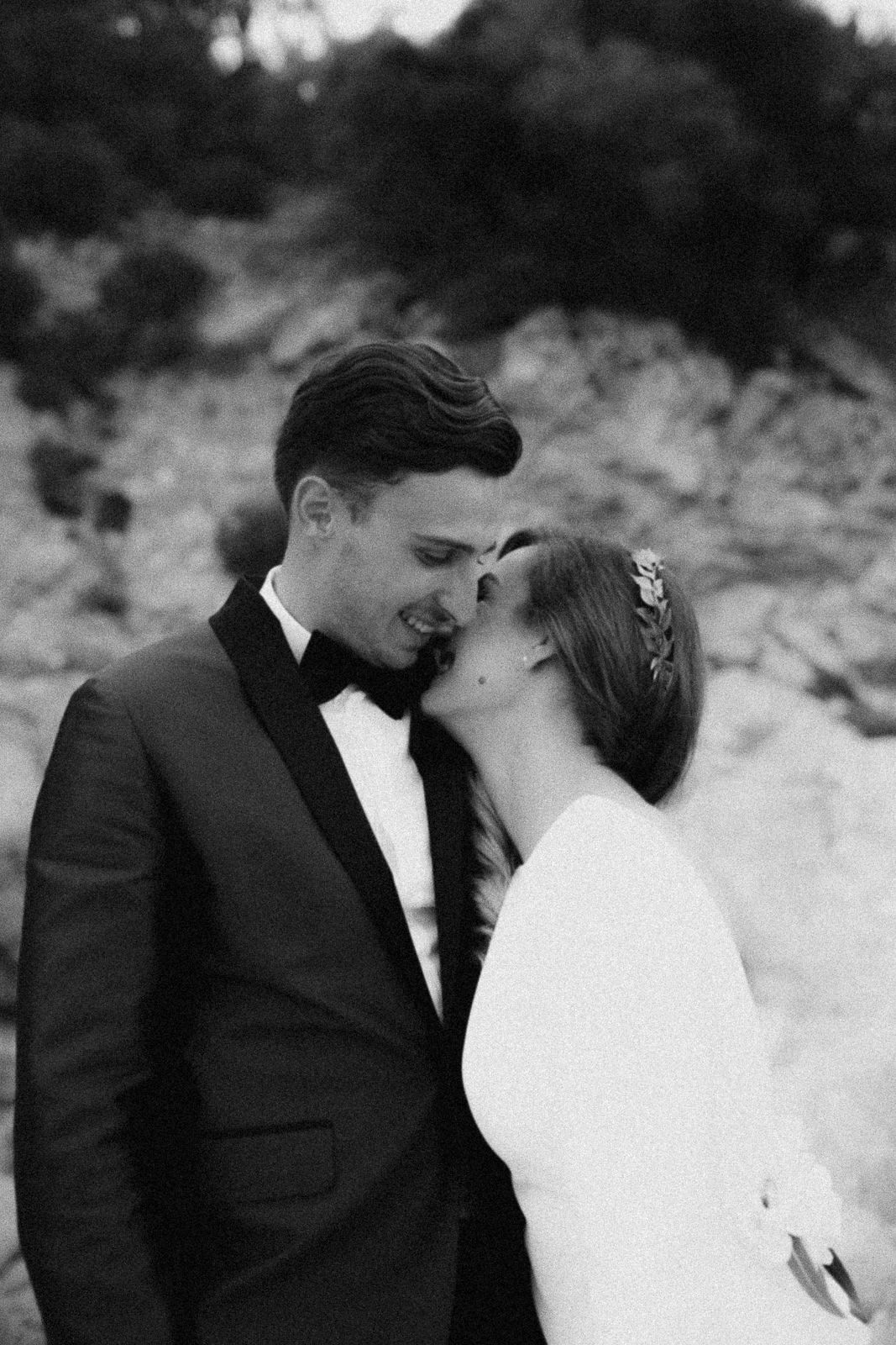 A romantic black and white wedding portrait shows a tender moment between a couple in formal attire outdoors.