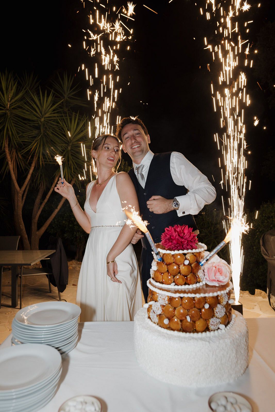 Wedding couple stands behind their elegant white wedding cake with sparklers creating a magical nighttime display.
