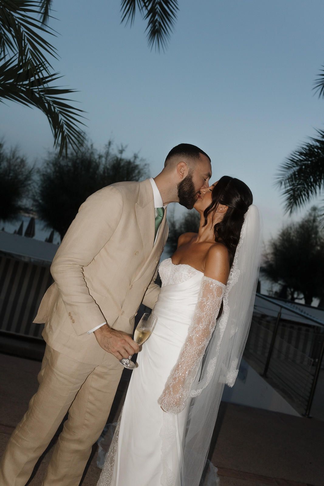 A romantic couple in beige suit and white wedding dress share a kiss at dusk under palm trees.