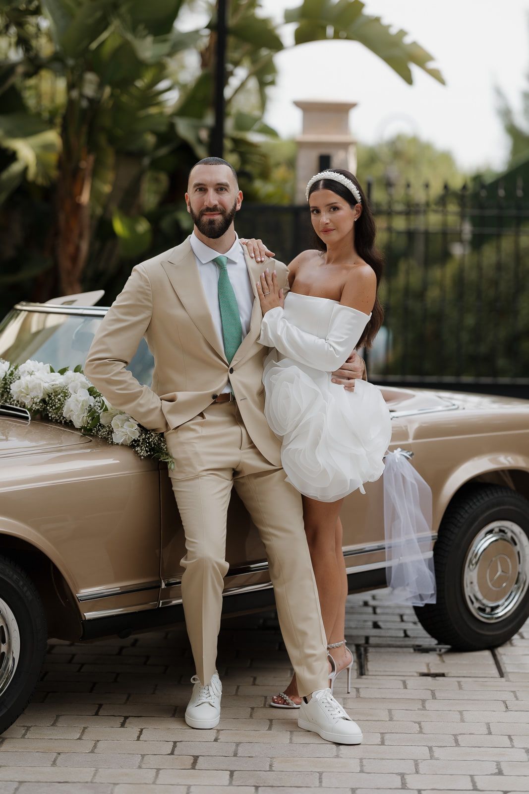 A newlywed couple poses playfully by a vintage champagne-colored car on a stone paved driveway.