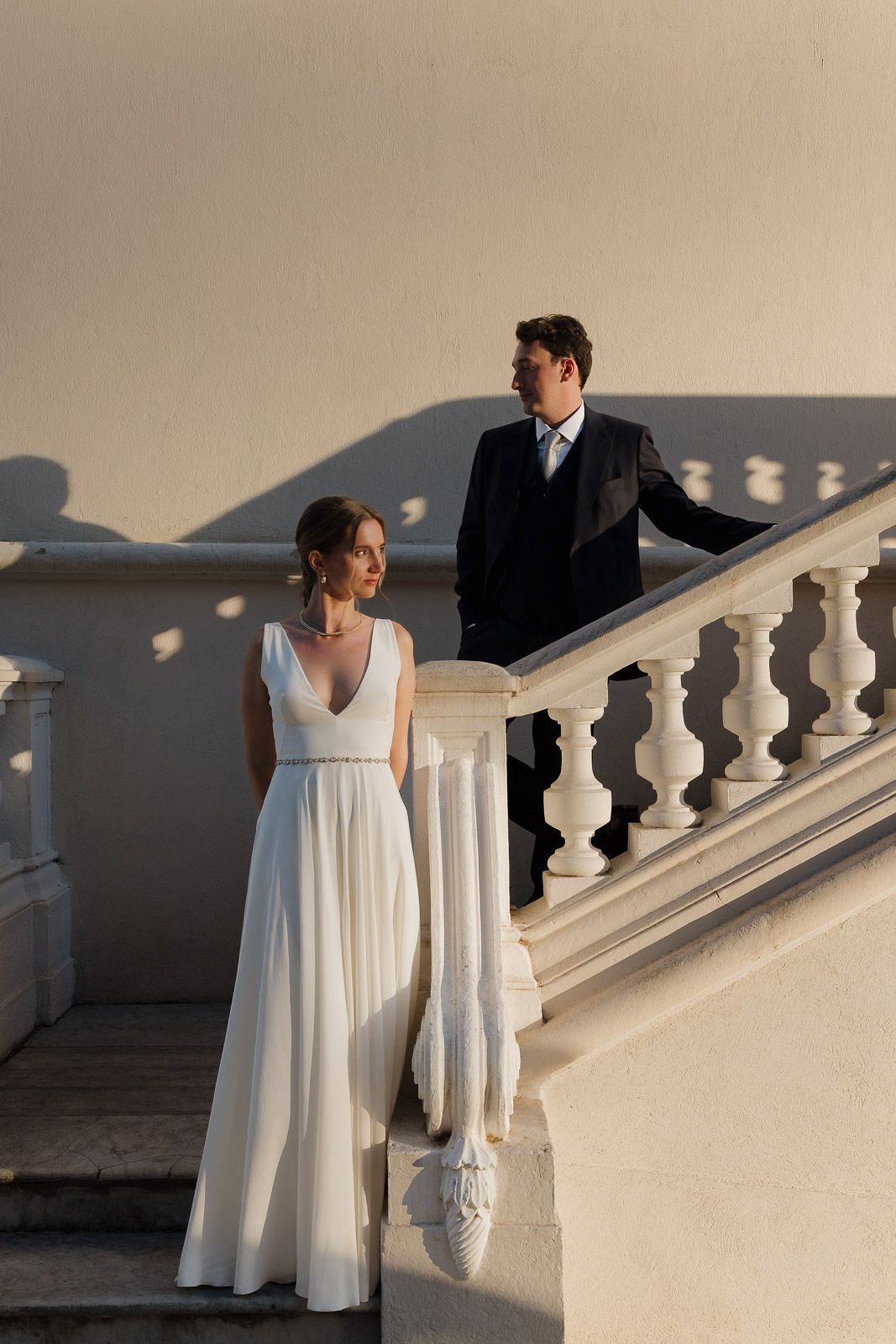 A bride in a flowing white dress and groom in dark suit stand on elegant white stone stairs at sunset.
