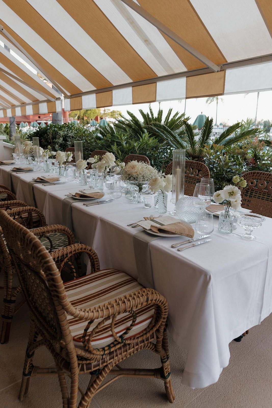 Elegant wedding reception setup with long white-clothed tables and rattan chairs under a striped awning.
