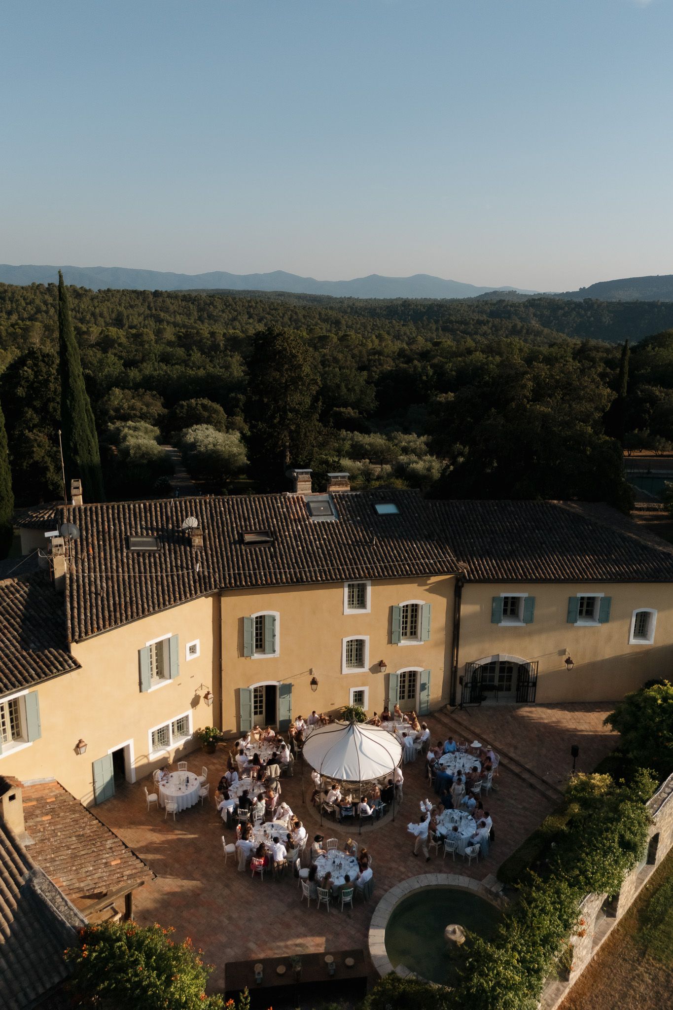 Aerial view of an elegant French countryside wedding reception in a golden villa courtyard, surrounded by lush forests and mountains.