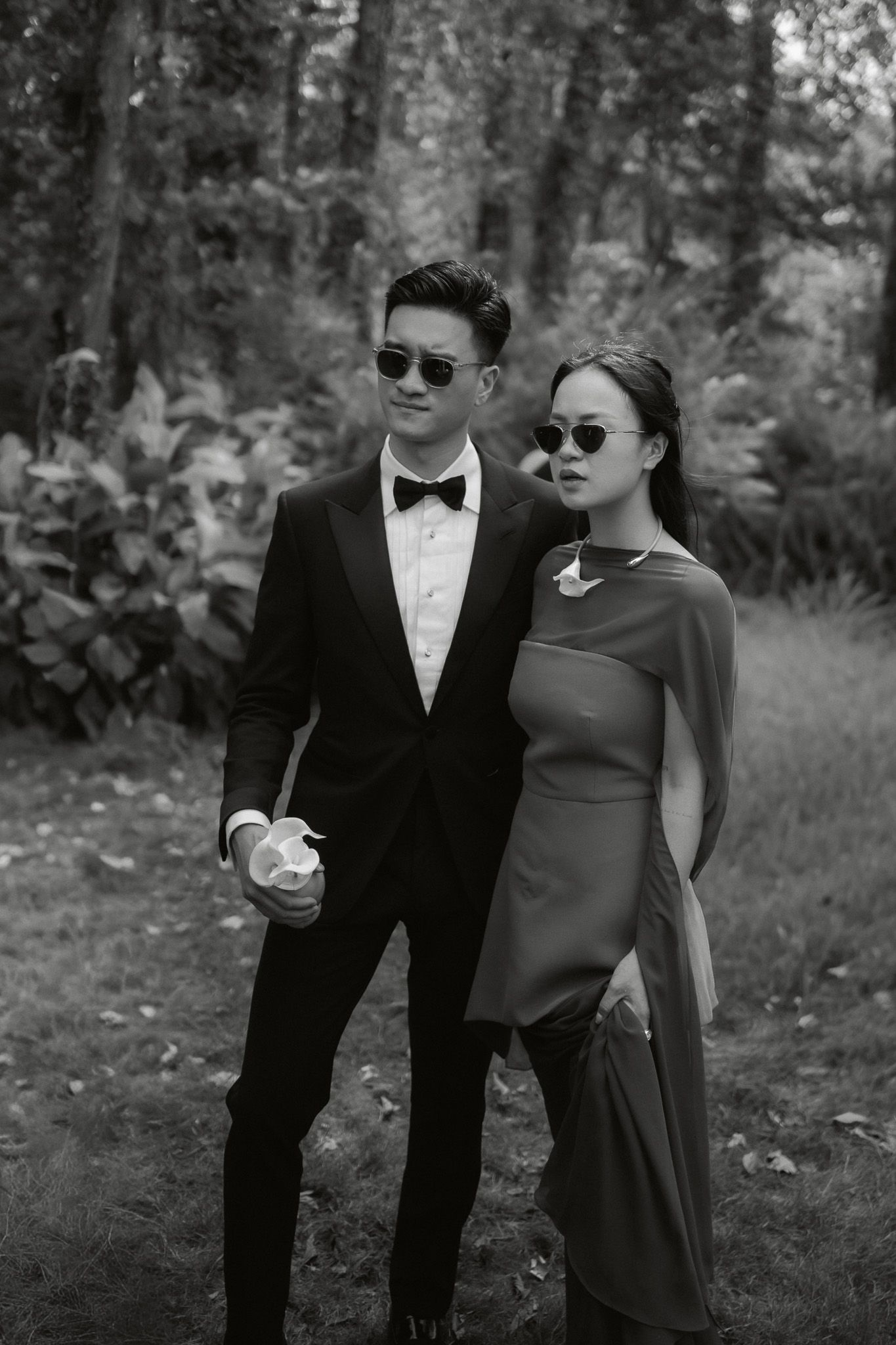 Elegant couple in black and white, man in tuxedo and woman in gown, posing in a lush garden setting.