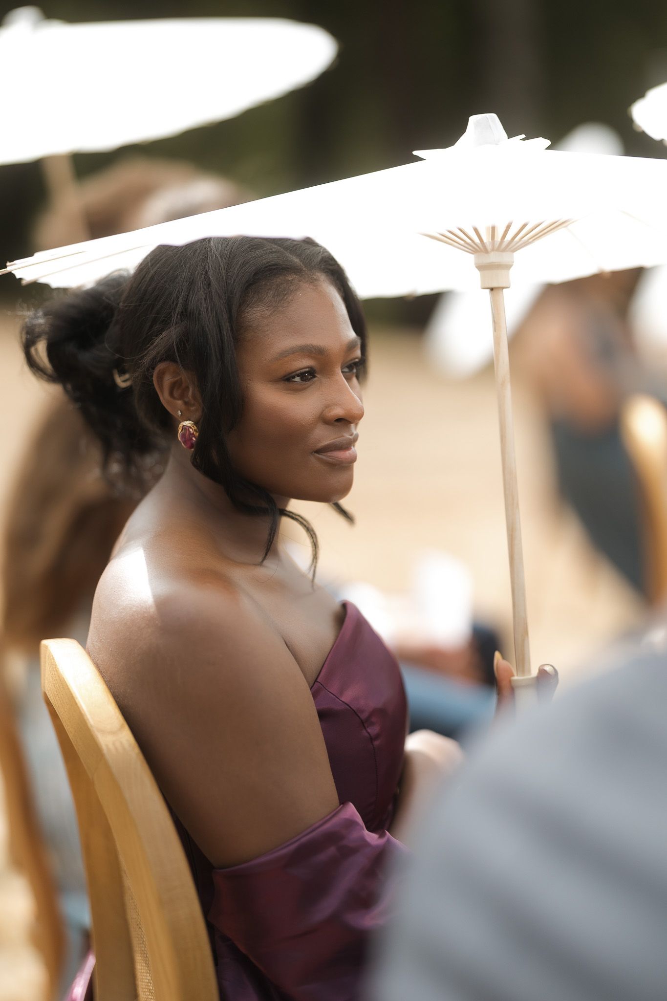 Elegant woman in purple strapless gown seated outdoors under white parasol at formal event.