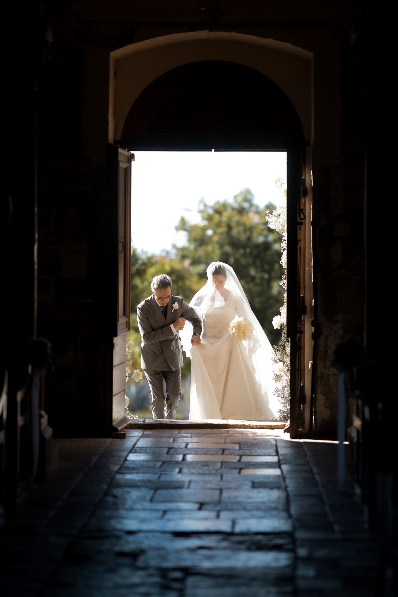 Bride in white gown and veil enters church doorway with escort, backlit by natural sunlight.