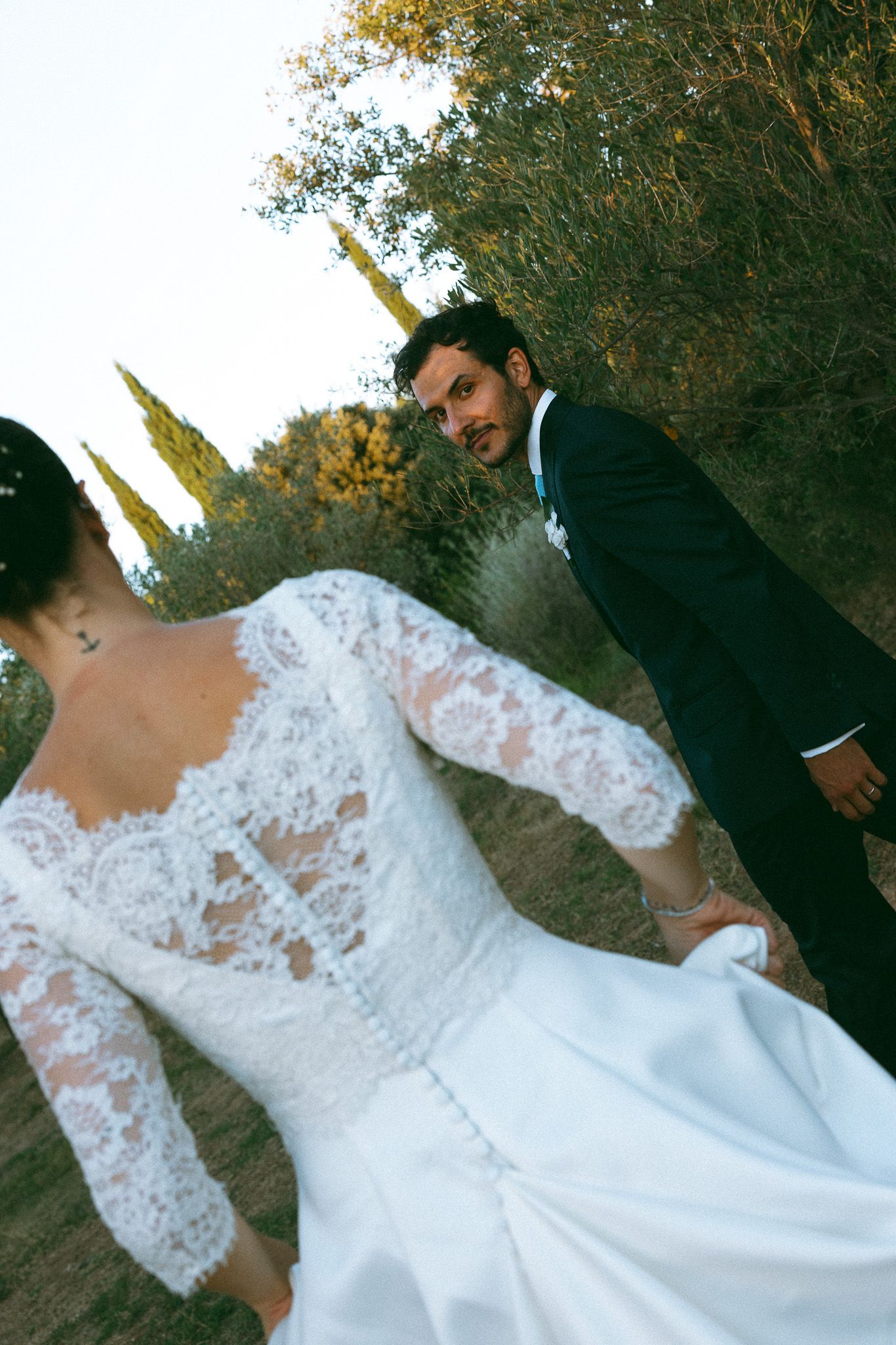 Bride in lace long-sleeve wedding gown holds groom's hand outdoors, as he looks back smiling in a black suit.