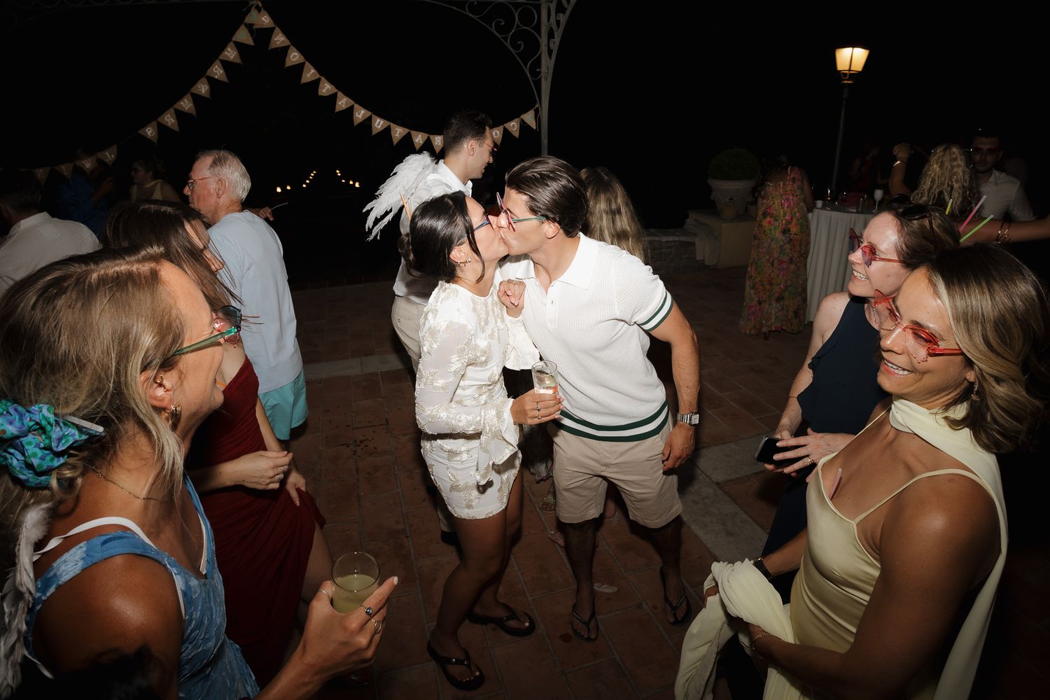Couple sharing a kiss at outdoor wedding reception at night surrounded by smiling guests.